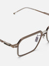 Close-up of A. SOCIETY x Universal Works Kowloon Eyewear in brown/honey on a white surface. The glasses have a unique rectangular frame and clear nose pads.