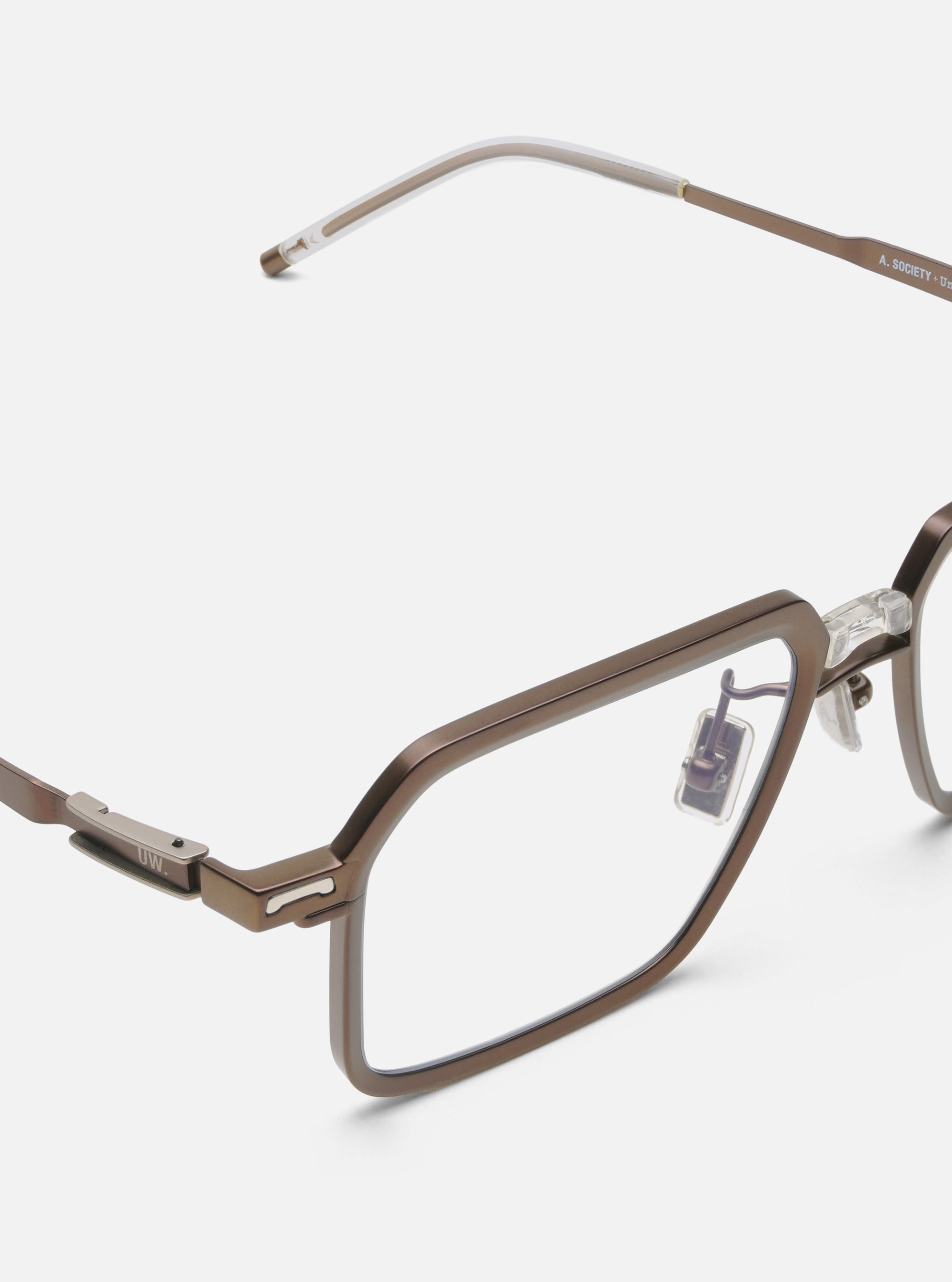 Close-up of A. SOCIETY x Universal Works Kowloon Eyewear in brown/honey on a white surface. The glasses have a unique rectangular frame and clear nose pads.