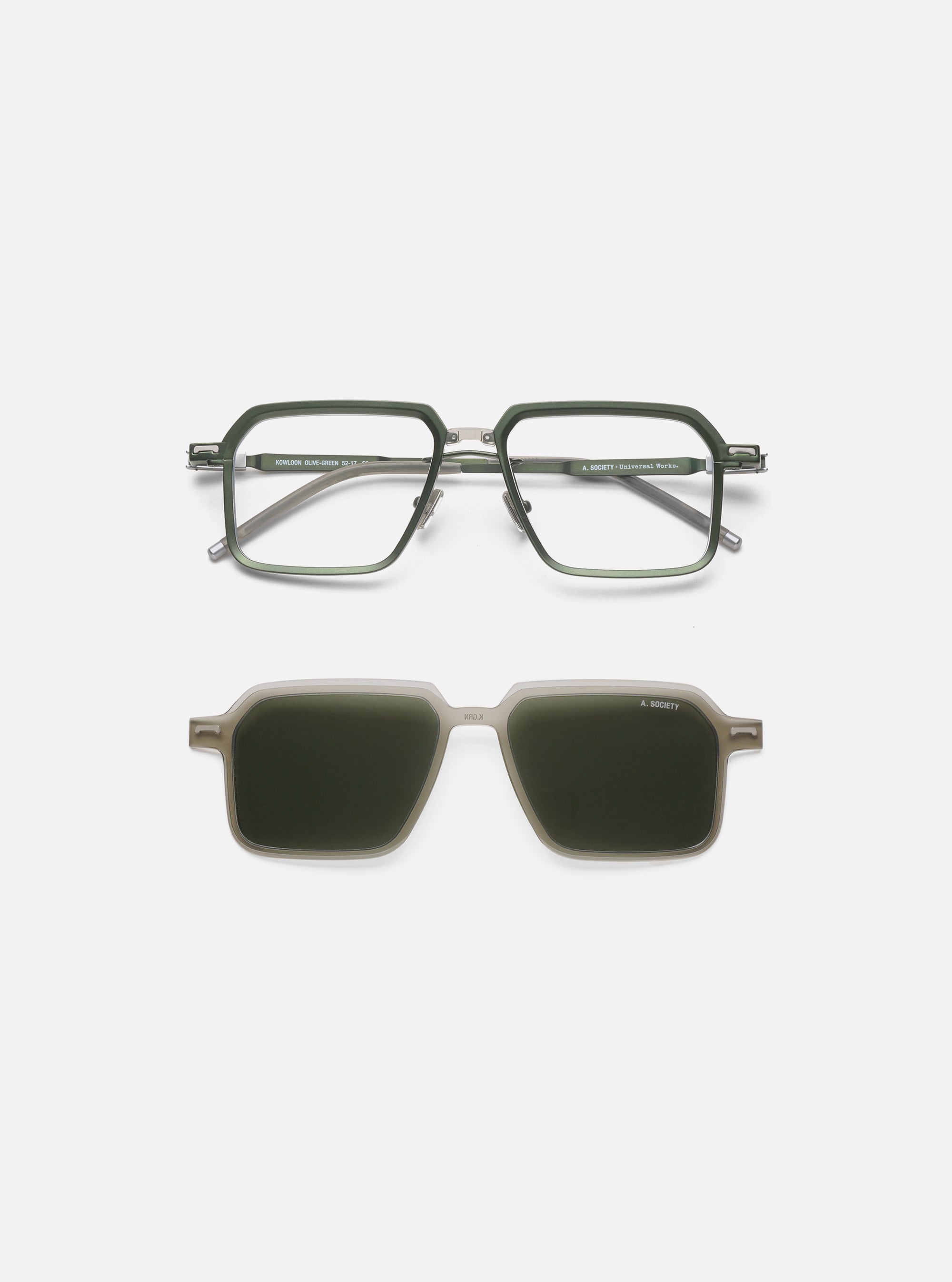 A. SOCIETY x Universal Works Olive/Green Kowloon Eyewear on a white surface. The glasses have an olive green frame and clear lenses, with a clip-on sunglass attachment.