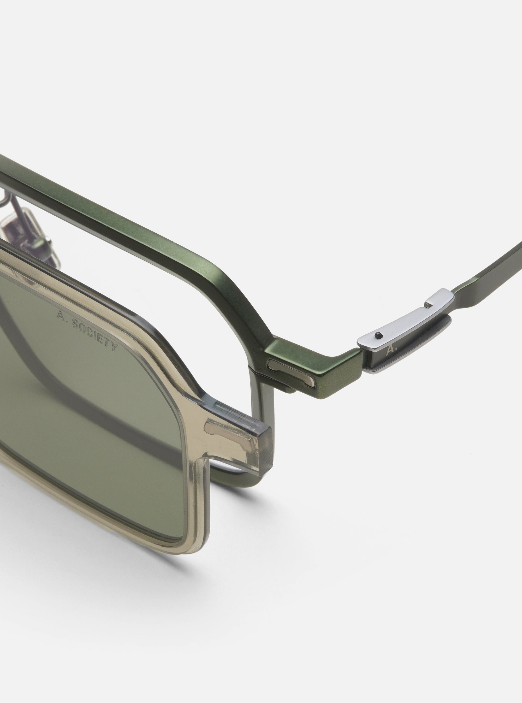 Close-up of A. SOCIETY x Universal Works Olive/Green Kowloon Eyewear on a white surface. Features olive green frames and silver accents. Stylish and modern eyewear.