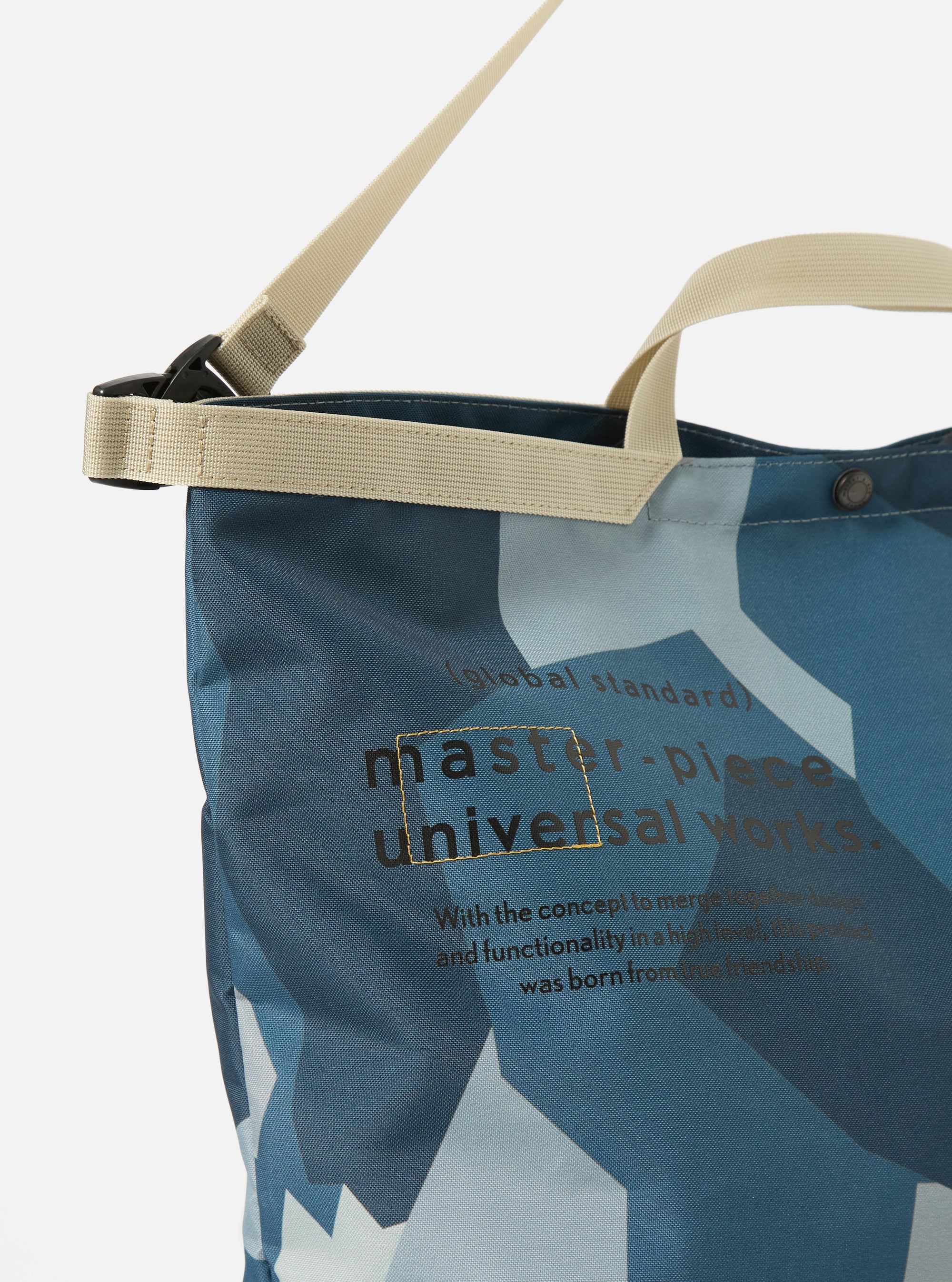 Close-up of the master-piece x Universal Works Blue Camo Recycled Tech Canvas Large Tote Bag. Features blue camo print, beige straps, and text on a white background.
