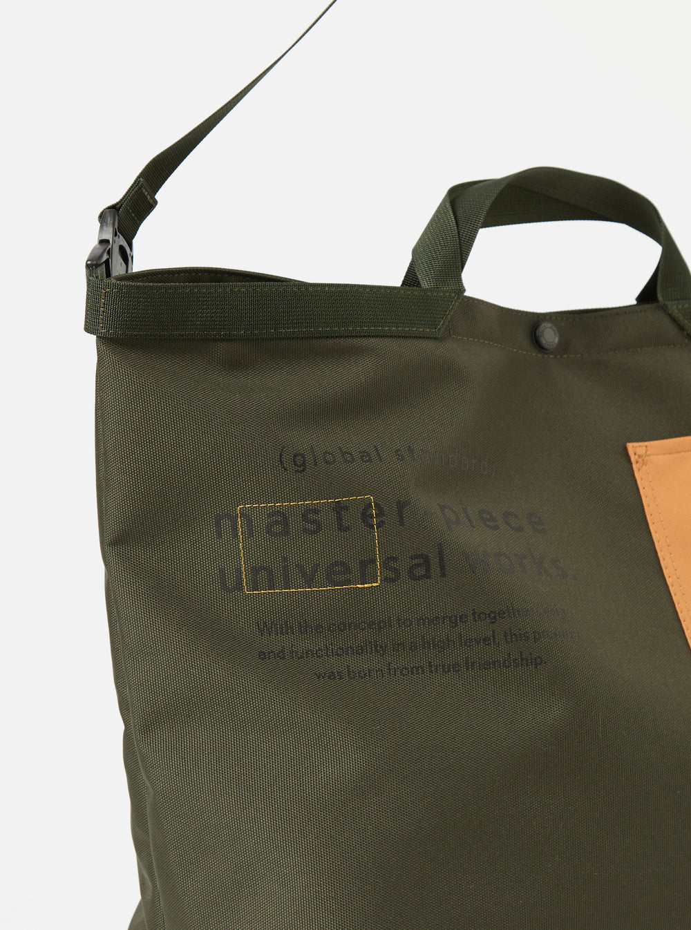 Close-up of the master-piece x Universal Works Olive Recycled Tech Canvas Large Tote Bag. The olive green bag features black lettering and a tan patch. Shot on a white background.