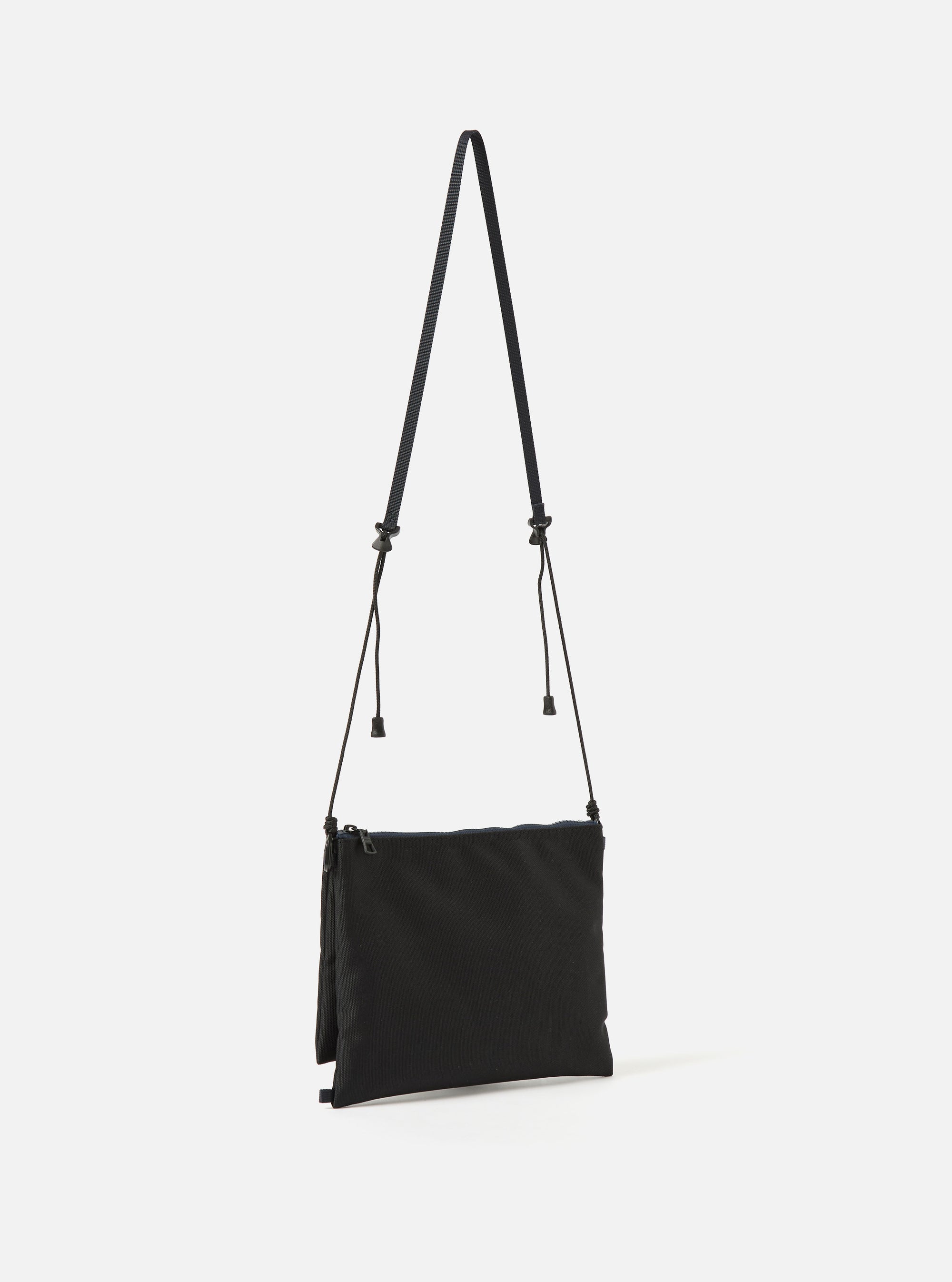 master-piece x Universal Works Black Recycled Tech Canvas Sacoche on a clean white background. The black bag hangs by a thin strap. The material looks durable and modern.