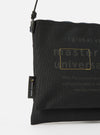 Close-up of the master-piece x Universal Works Black Recycled Tech Canvas Sacoche on a white background. The black canvas bag features subtle branding details.