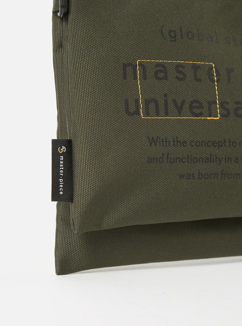 Close-up of the master-piece x Universal Works Olive Recycled Tech Canvas Sacoche on a white background. The olive canvas features printed text and a small brand tag.
