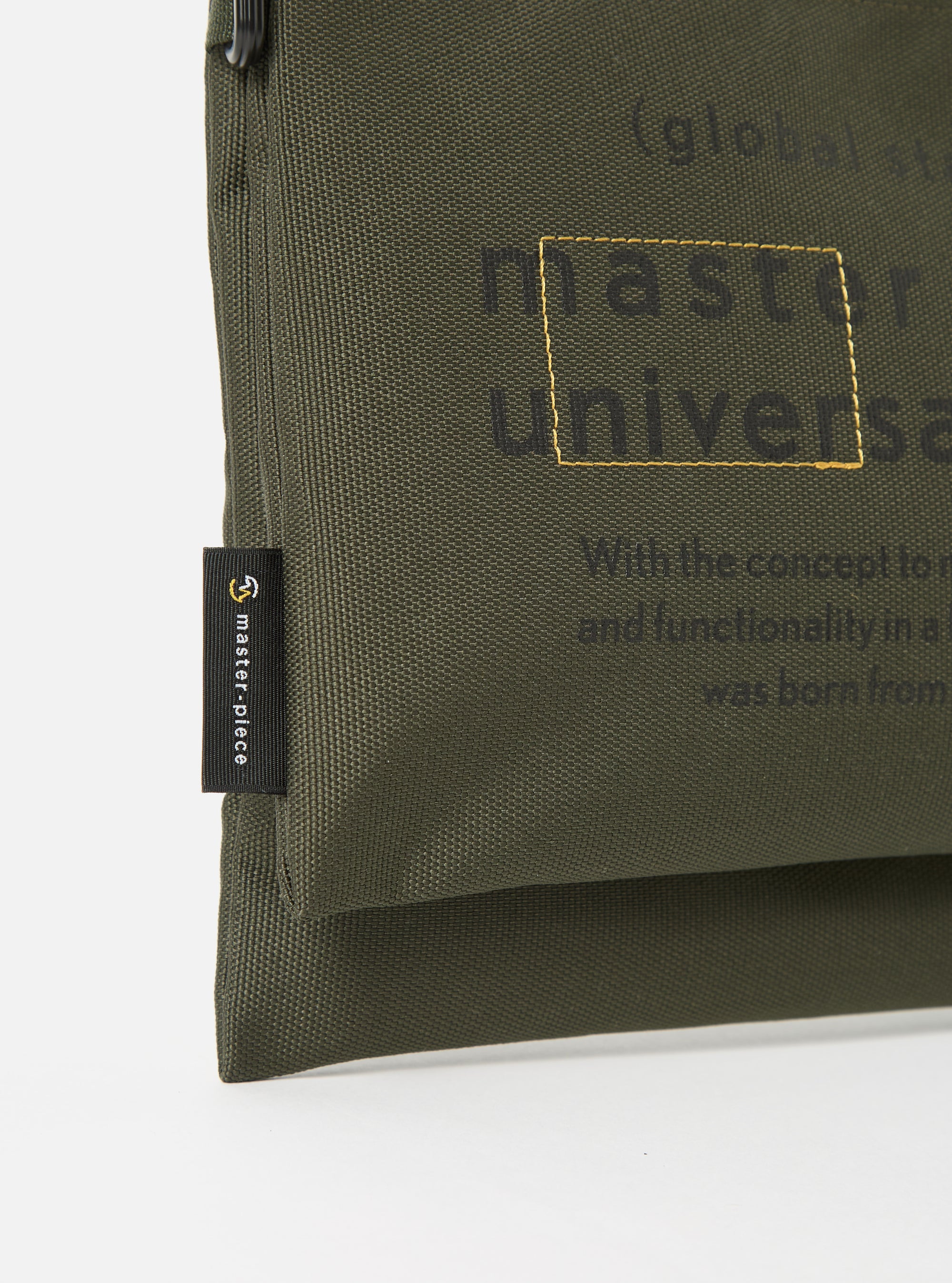 Close-up of the master-piece x Universal Works Olive Recycled Tech Canvas Sacoche on a white background. The olive canvas features printed text and a small brand tag.