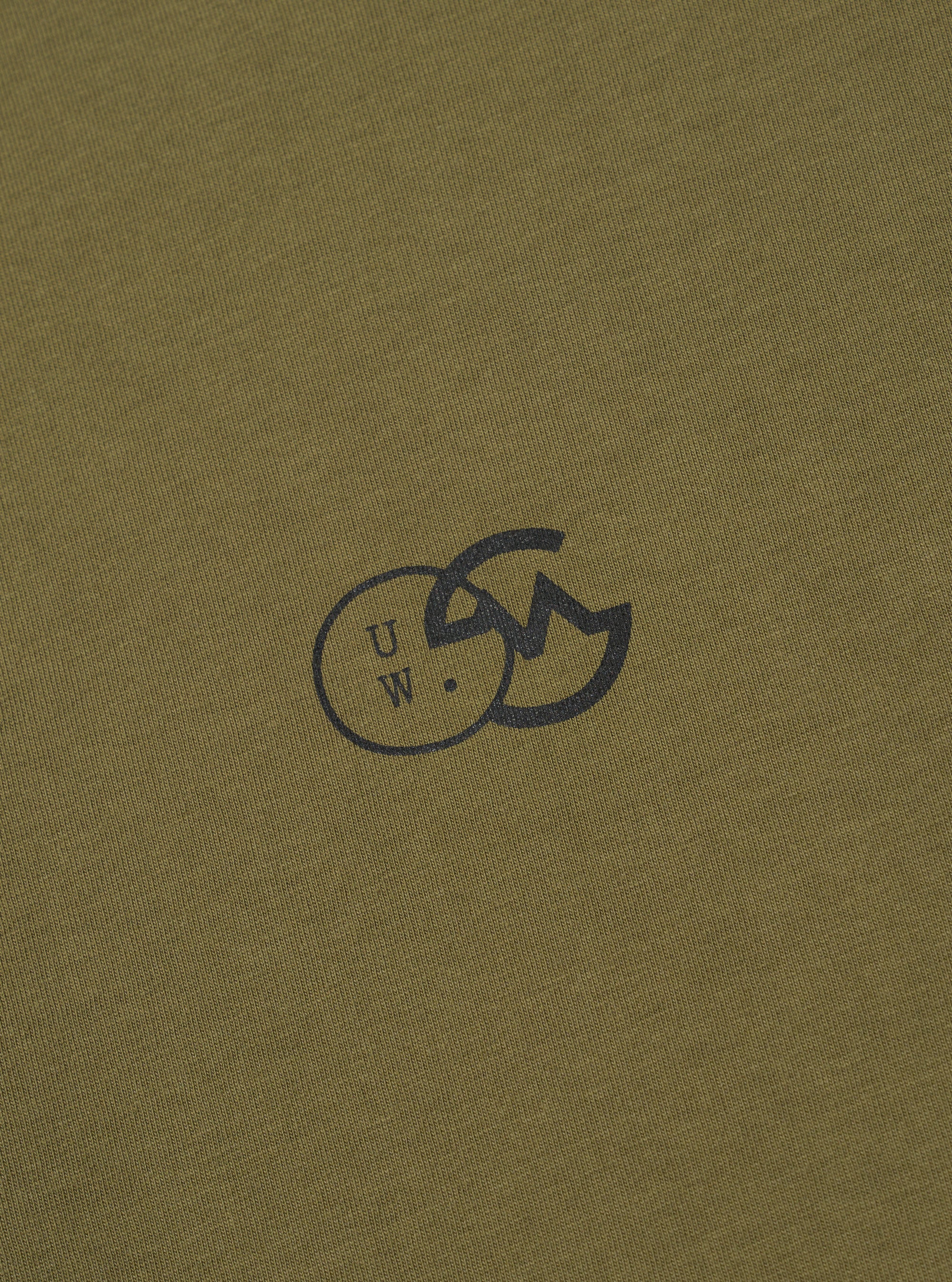 The master-piece x Universal Works Olive Single Jersey Print Tee is shown from the back as a flat lay against a white background. The image highlights the large, detailed graphic print on the back of the olive green t-shirt, which includes text and various illustrative elements.