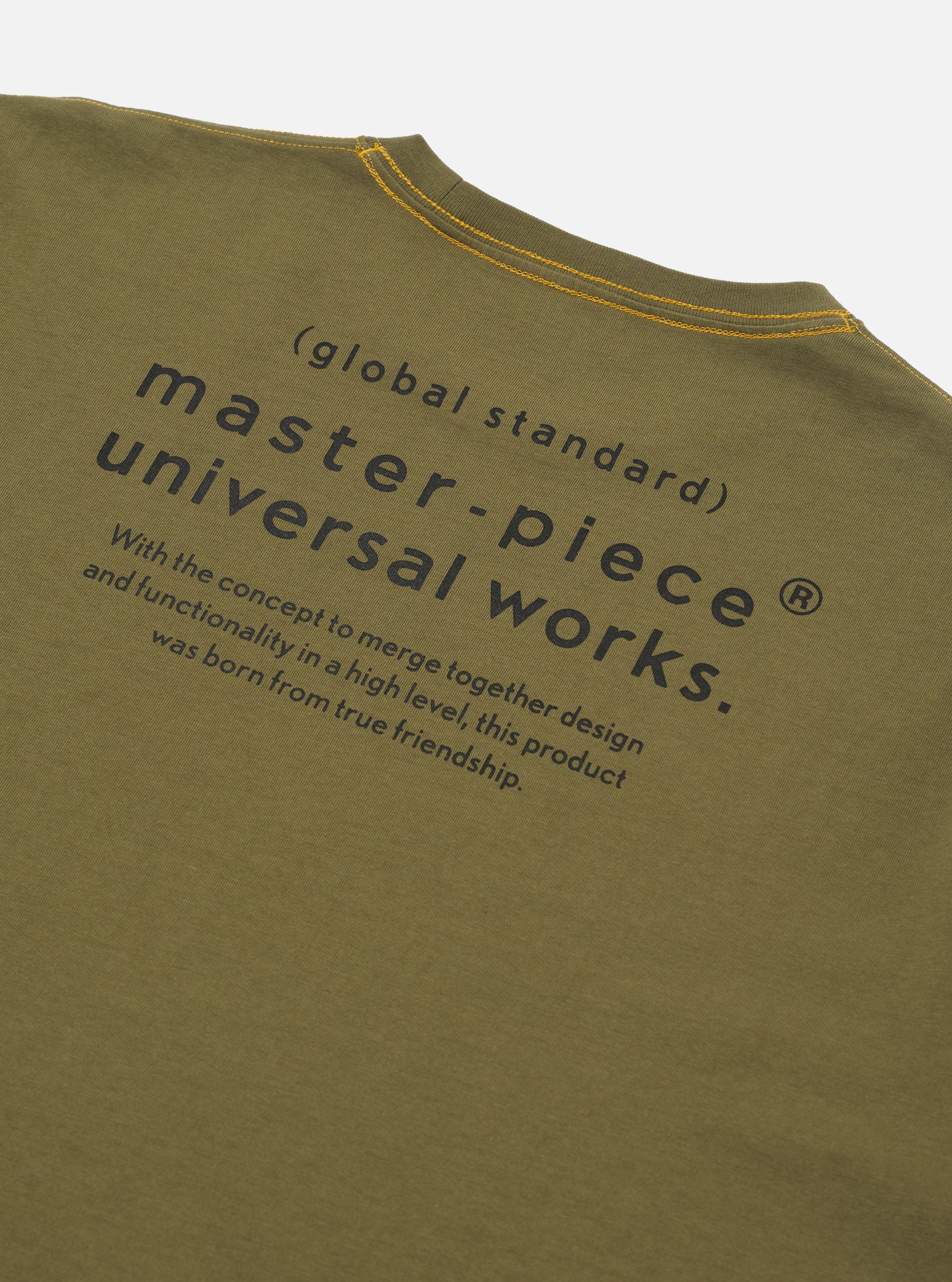 The master-piece x Universal Works Olive Single Jersey Print Tee is shown as a flat lay against a white background. The image displays a close-up detail of the garment, focusing on the back neck area and the internal branding labels. A blue master-piece brand tag is visible next to a white Universal Works label. The olive green cotton jersey fabric is prominent.