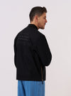 The master-piece x Universal Works Black Peached Poplin Newark Jacket is worn by a model standing against a plain white background. The image shows the model from the back, highlighting the clean lines of the black jacket, its ribbed hem, and the collaboration branding near the neckline. The jacket is styled over a light-colored shirt and olive trousers.