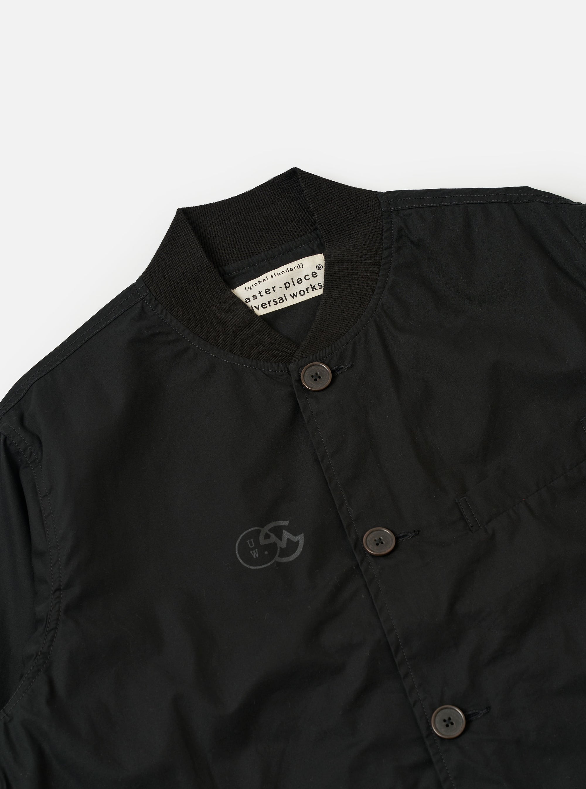 The master-piece x Universal Works Black Peached Poplin Newark Jacket is shown as a flat lay against a plain white background. The jacket features a rounded collar, a central button-down front, and two large patch pockets at the waist. The black peached poplin fabric has a smooth, matte finish.