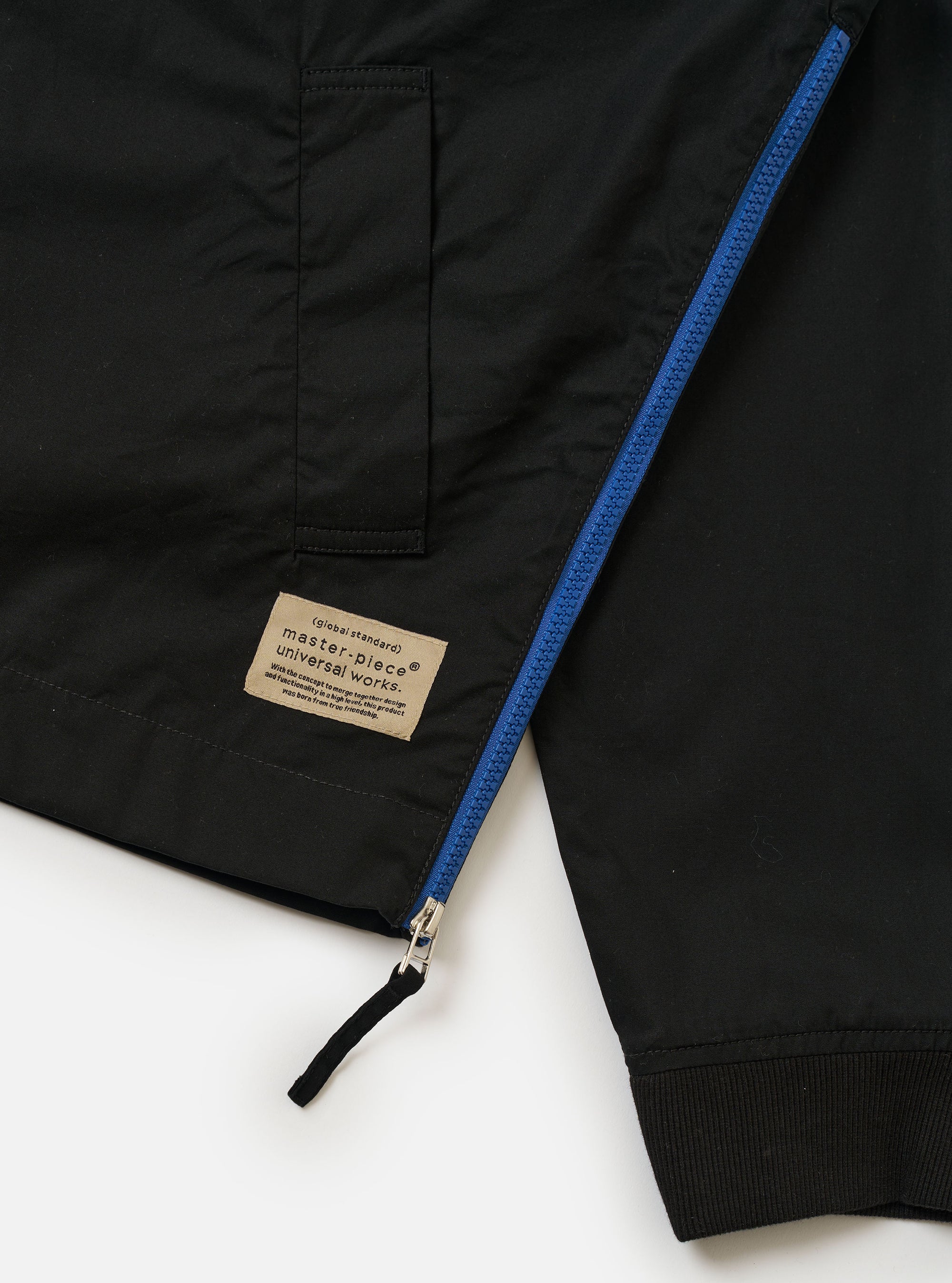 The master-piece x Universal Works Black Peached Poplin Newark Jacket is shown from a rear three-quarter angle as a flat lay against a plain white background. The image highlights the smooth black cotton fabric, the ribbed collar and cuffs, and a small collaborative brand patch near the hem.