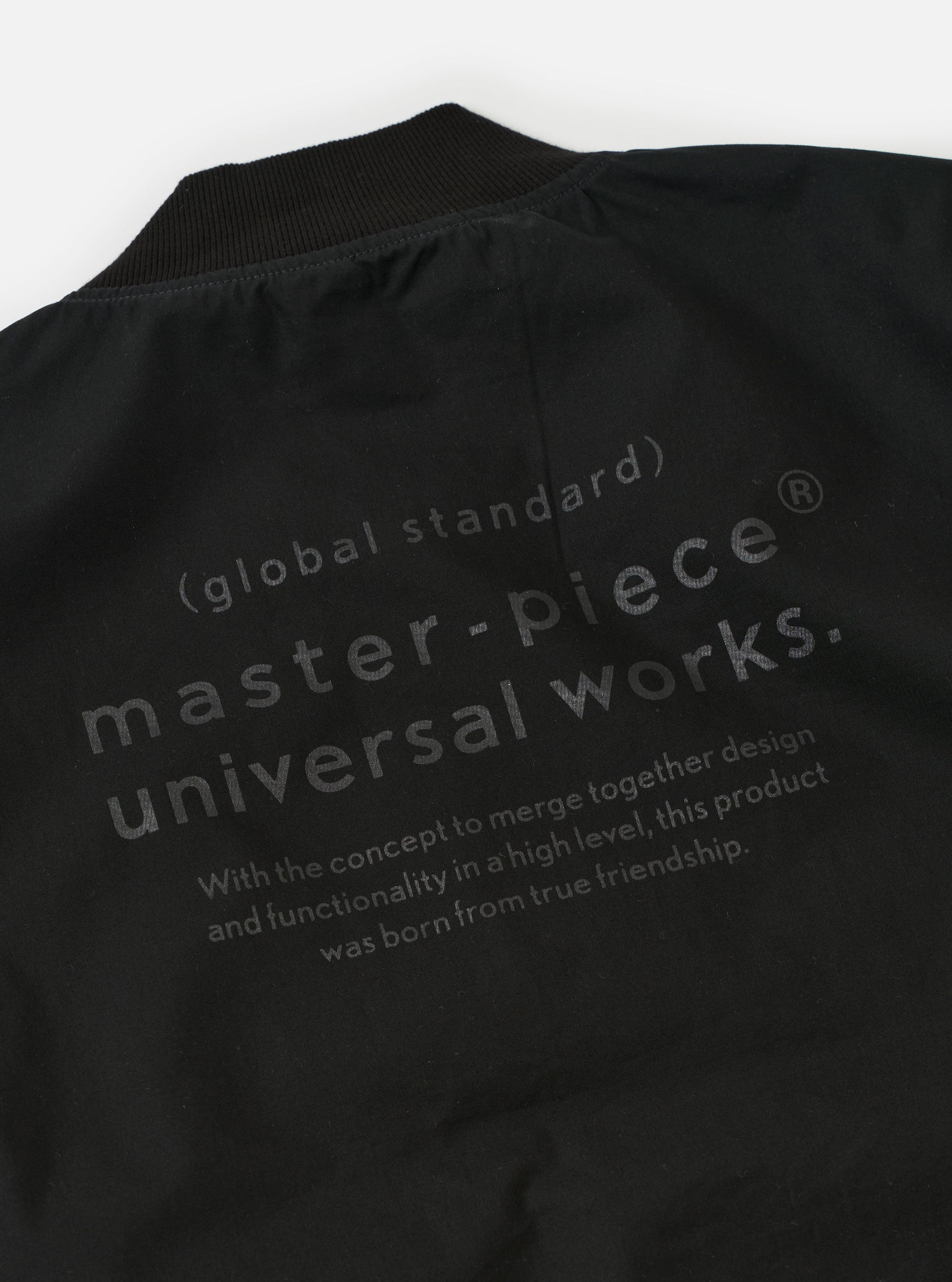 The master-piece x Universal Works Black Peached Poplin Newark Jacket is shown in a detailed close-up shot as a flat lay against a plain white background. The image focuses on the interior branding and construction, highlighting the master-piece x Universal Works dual-branded label and a white care instruction patch sewn onto the black fabric near a pocket.