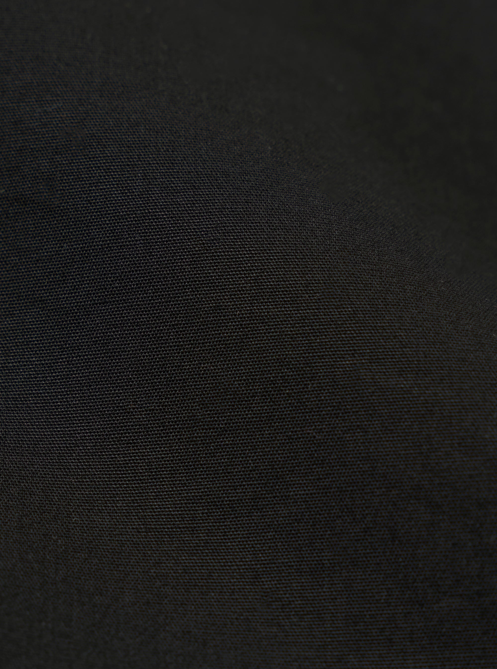 A close-up fabric swatch of the master-piece x Universal Works Black Peached Poplin Newark Jacket is shown against a plain white background. The image highlights the smooth, matte texture of the black peached poplin material and its fine weave.