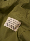 The master-piece x Universal Works Olive Recycled Poly Tech Tech Overshirt is shown in a close-up detail shot as a flat lay against a white background. The image focuses on the elasticated drawcord at the hem and the concealed side seam pocket with a tonal zip. The smooth, utilitarian texture of the olive green recycled polytech fabric is clearly visible.