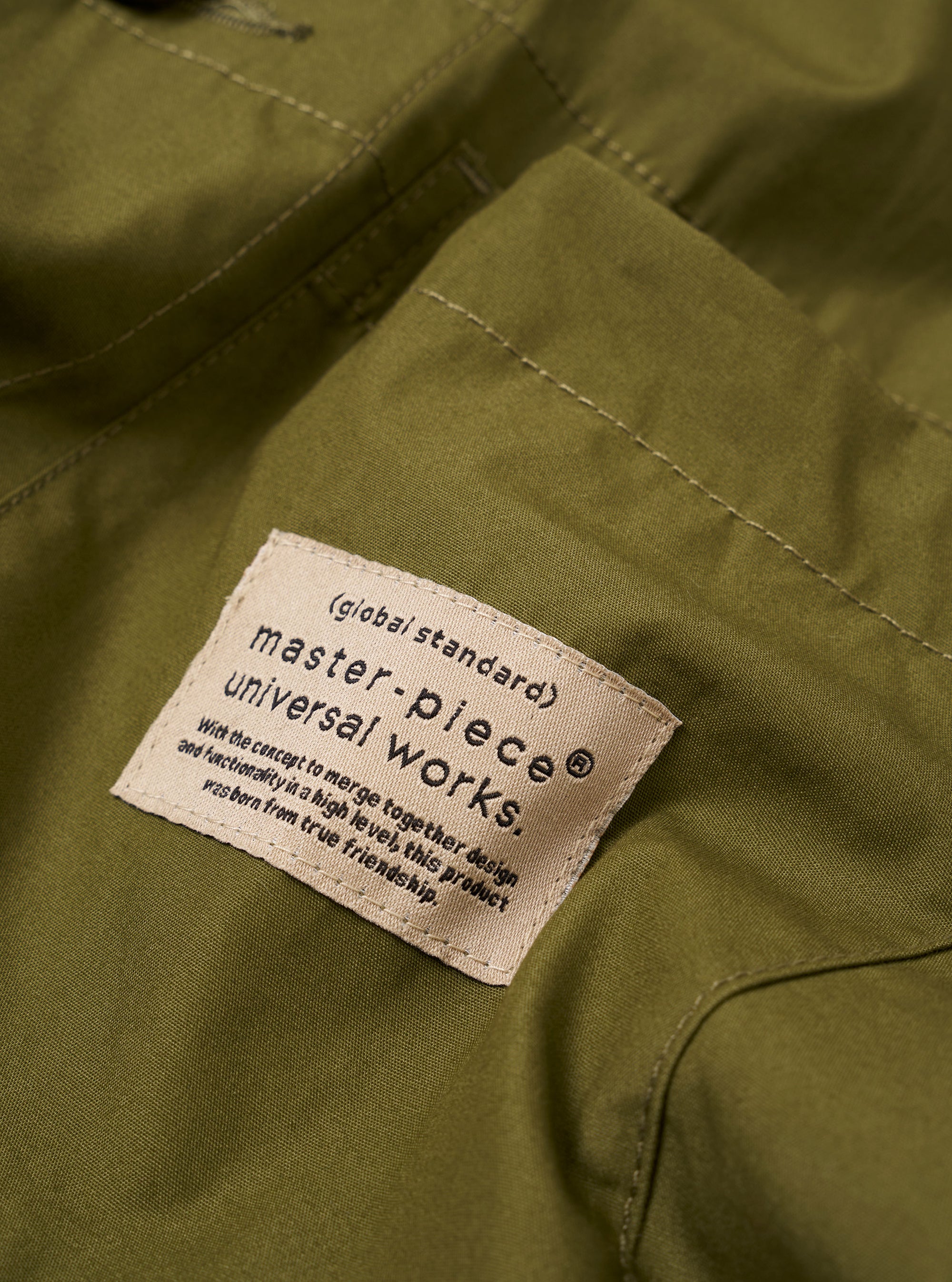 The master-piece x Universal Works Olive Recycled Poly Tech Tech Overshirt is shown in a close-up detail shot as a flat lay against a white background. The image focuses on the elasticated drawcord at the hem and the concealed side seam pocket with a tonal zip. The smooth, utilitarian texture of the olive green recycled polytech fabric is clearly visible.
