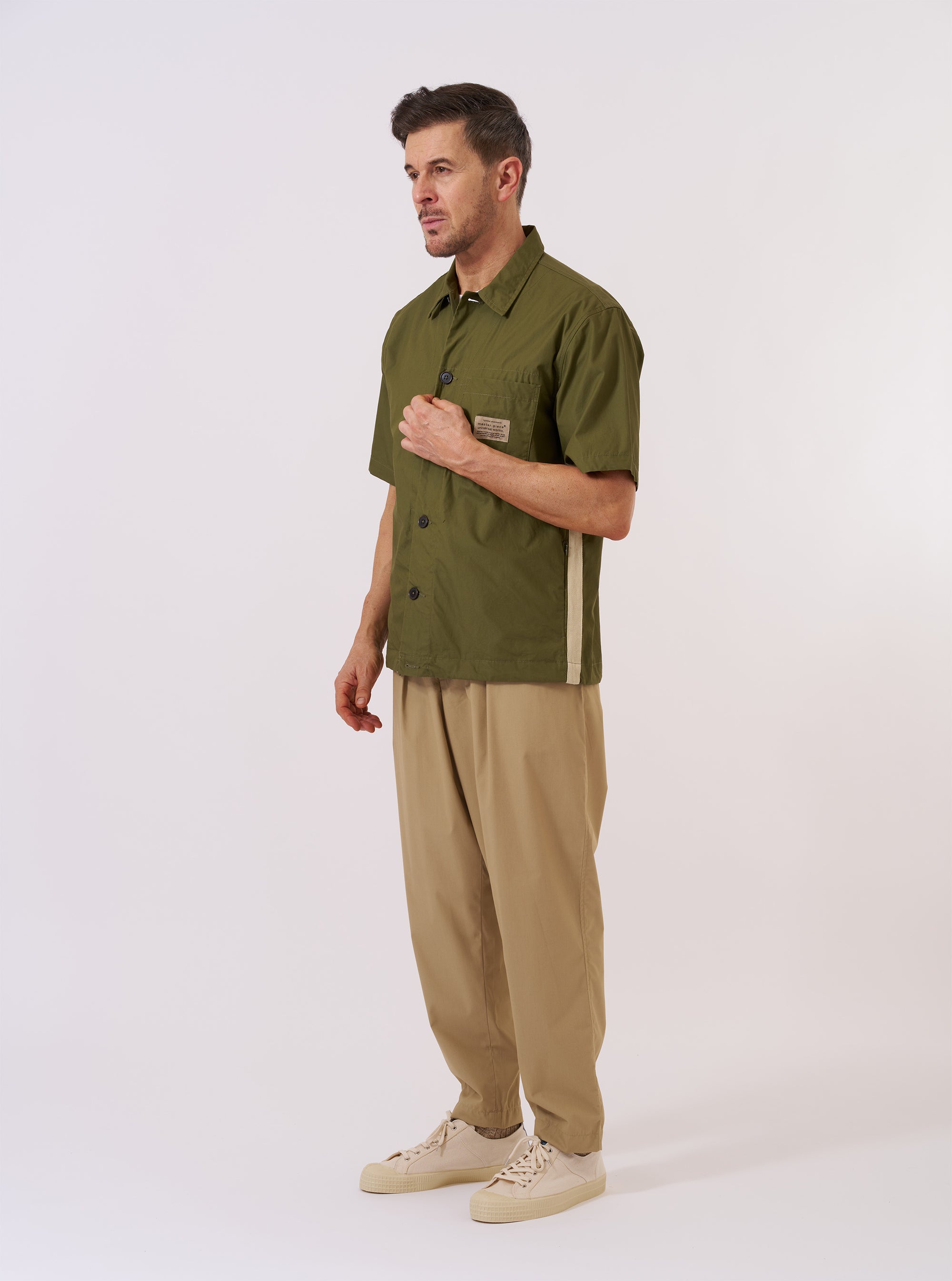 The master-piece x Universal Works Olive Recycled Poly Tech Tech Overshirt is shown as a flat lay against a plain white background. This short-sleeve overshirt features a one-piece collar, a full button-through front, and a single patch pocket on the left chest. The olive green fabric has a clean, utilitarian appearance.