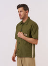 A model wears the master-piece x Universal Works Olive Recycled Poly Tech Tech Overshirt against a plain white background. The olive green overshirt features short sleeves, a one-piece collar, a central button-down closure, and a single patch pocket on the left chest. The model is shown from the front, with the garment displaying a clean, technical finish and a straight hem.
