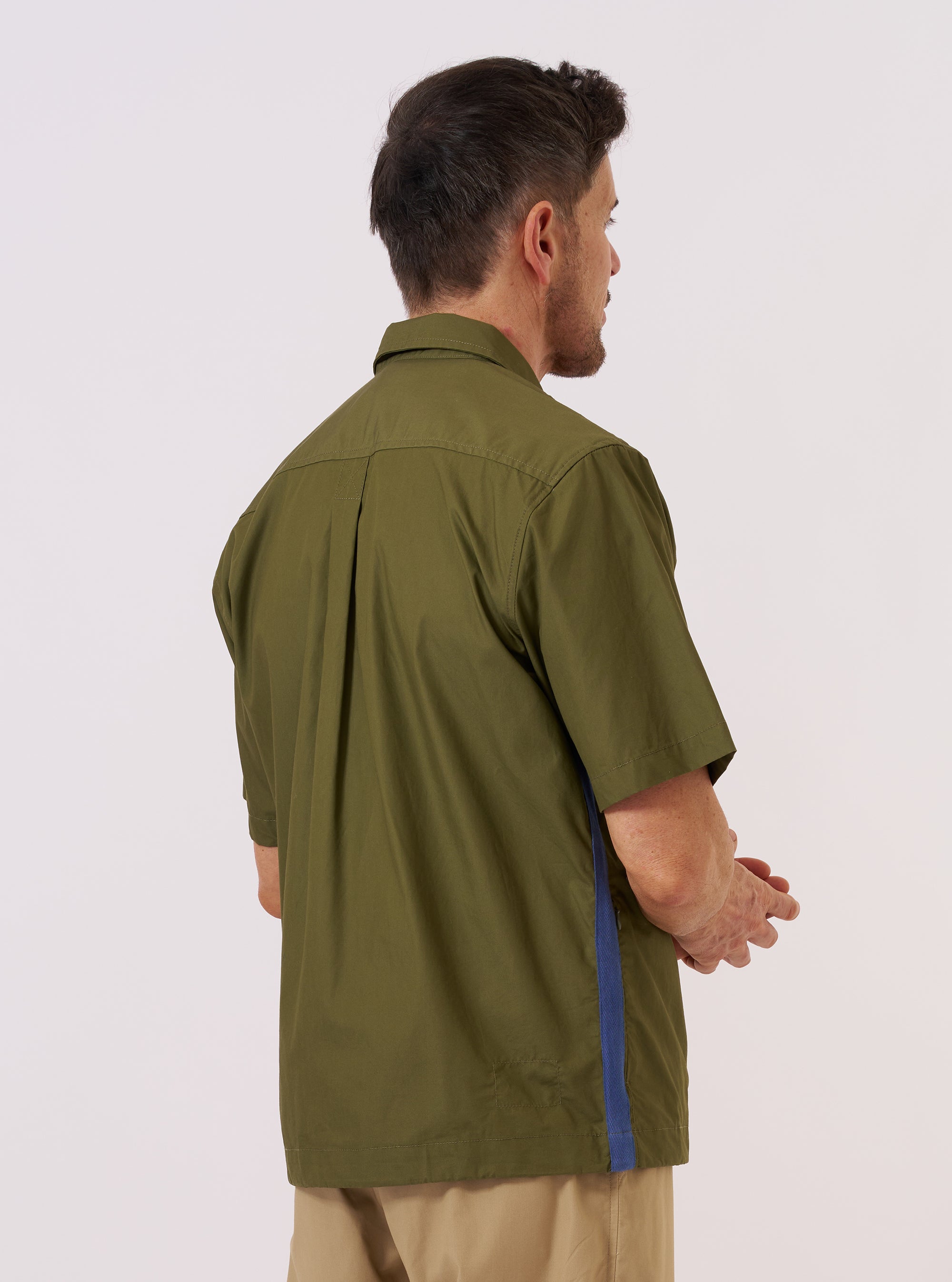 A model is wearing the master-piece x Universal Works Olive Recycled Poly Tech Tech Overshirt against a plain white background. Shown from a rear three-quarter angle, the image highlights the overshirt's boxy fit, back yoke with a central box pleat, and a straight hem. The olive green recycled polytech fabric has a smooth, utilitarian finish. No mannequin is present in the image.