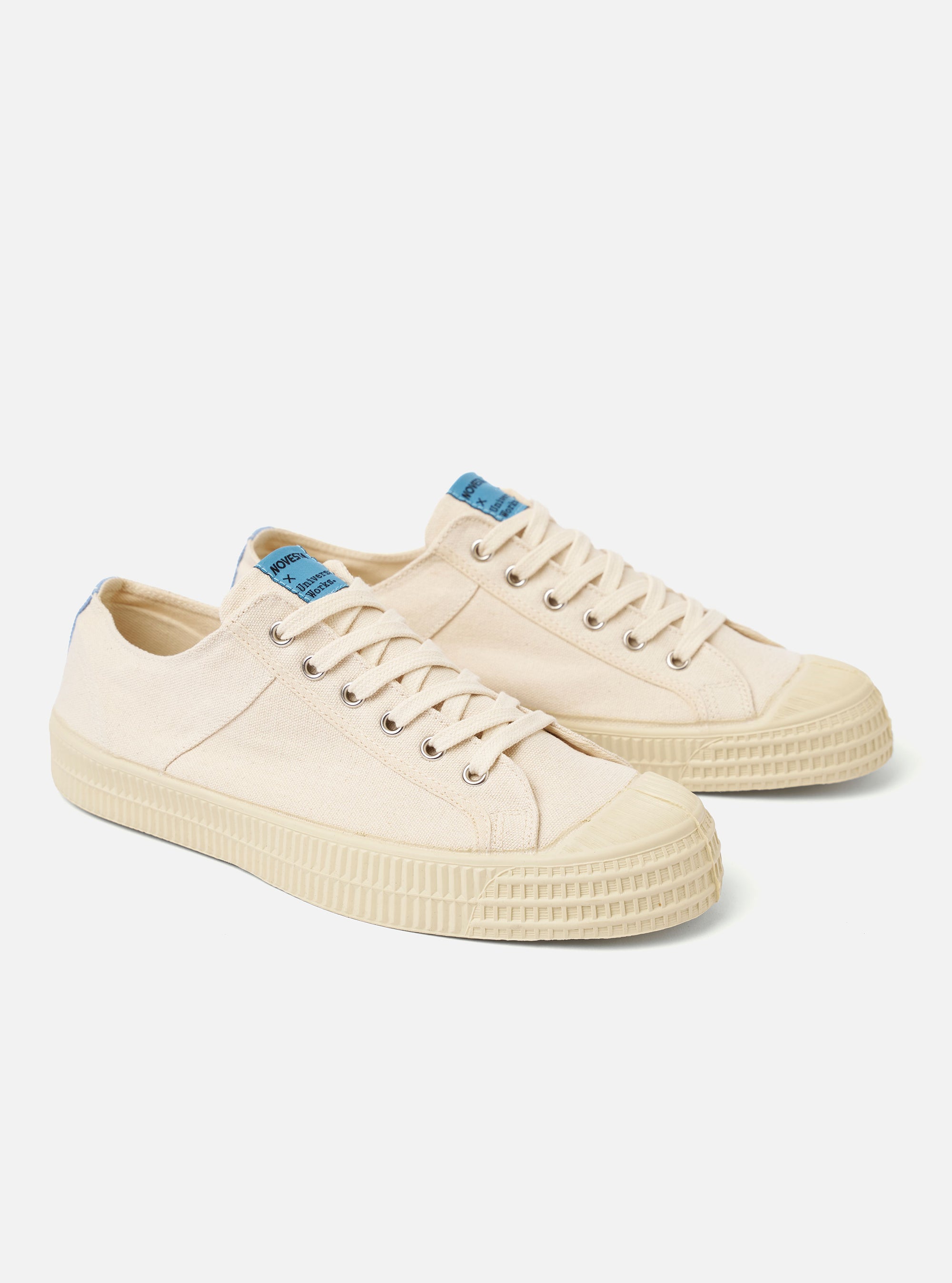 The Novesta for Universal Works Ecru/Pale Blue Canvas Star Master is shown from a side profile view against a white background. The low-top sneaker features an ecru cotton canvas upper with contrast stitching, metallic eyelets, and a signature chunky vulcanized rubber sole in ecru. A pale blue pull tab is visible on the heel, and a dual-branded woven tag is attached to the tongue.