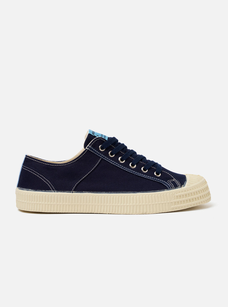 The Novesta for Universal Works Navy/Pale Blue Canvas Star Master is shown from a side profile against a plain white background. The low-top trainer features a navy cotton canvas upper with visible contrast stitching and silver metallic eyelets with navy laces. It has a prominent ecru rubber vulcanized sole with a textured finish and a matching ecru rubber toe cap. A pale blue fabric heel pull or flash is visible at the back of the shoe.