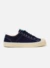 The Novesta for Universal Works Navy/Pale Blue Canvas Star Master is shown from a side profile against a plain white background. The low-top trainer features a navy cotton canvas upper with visible contrast stitching and silver metallic eyelets with navy laces. It has a prominent ecru rubber vulcanized sole with a textured finish and a matching ecru rubber toe cap. A pale blue fabric heel pull or flash is visible at the back of the shoe.