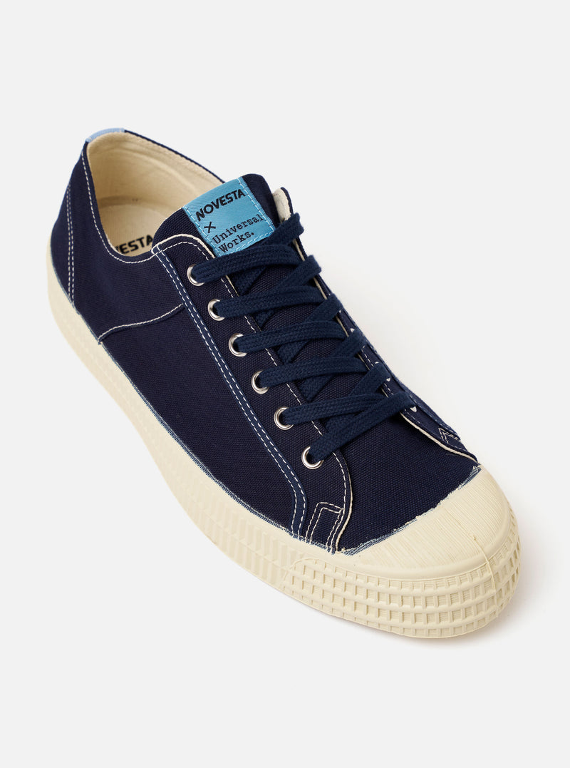 The Novesta for Universal Works Navy/Pale Blue Canvas Star Master sneaker is shown as a side profile shot against a white background. The shoe features a navy cotton canvas upper with contrast white stitching and silver metallic eyelets. It has a chunky ecru-colored natural rubber sole with a distinctive tire-like texture on the toe cap and heel bumper. A prominent pale blue pull tab is visible on the heel.