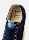 The Novesta for Universal Works Navy/Pale Blue Canvas Star Master is shown from a side profile against a white background. The low-top sneaker features a navy cotton canvas upper with contrast stitching, metallic eyelets, and a chunky ecru vulcanized rubber sole with a textured toe cap. A prominent pale blue pull tab is located at the heel.
