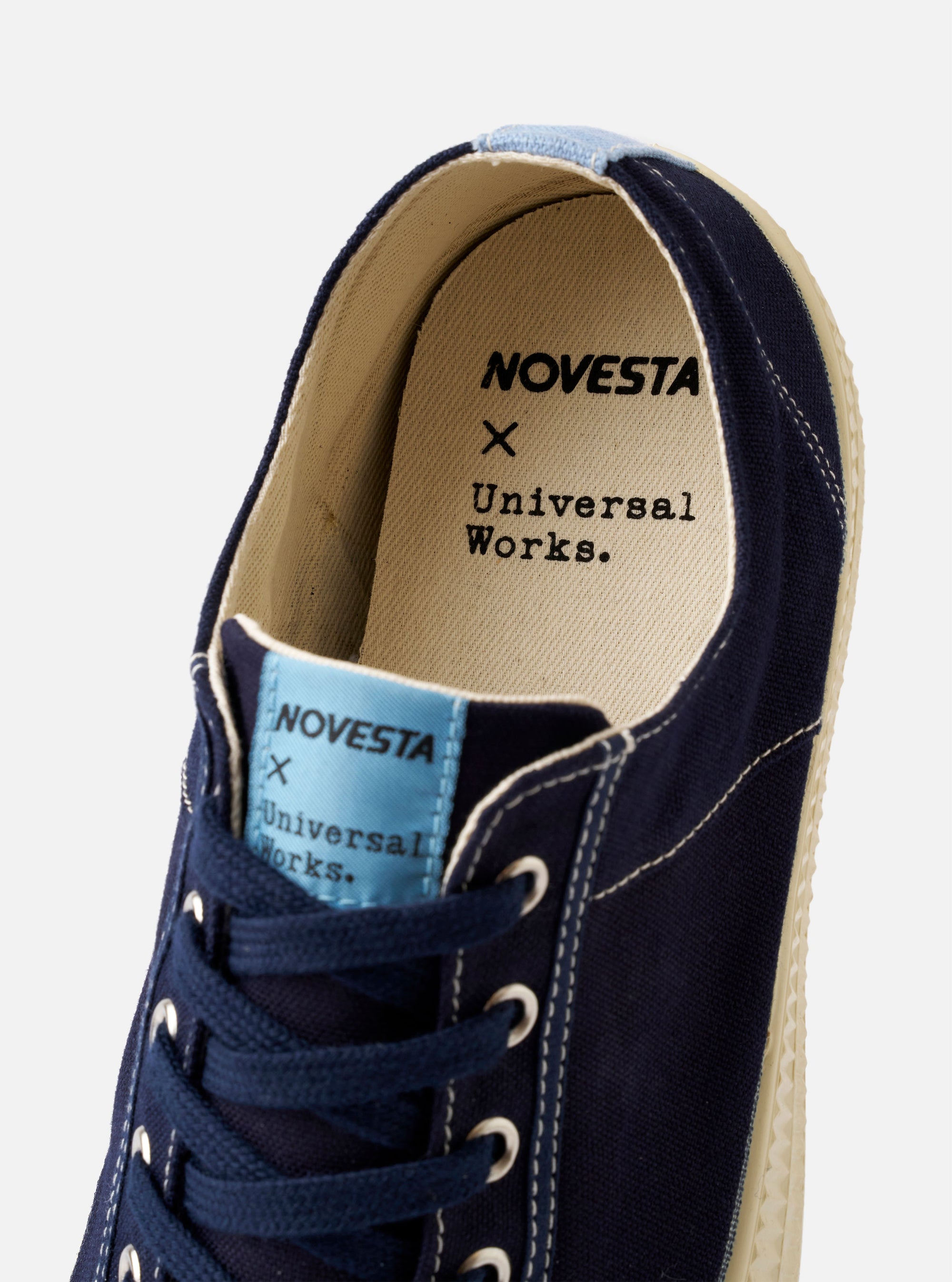 The Novesta for Universal Works Navy/Pale Blue Canvas Star Master is shown from a side profile against a white background. The low-top sneaker features a navy cotton canvas upper with contrast stitching, metallic eyelets, and a chunky ecru vulcanized rubber sole with a textured toe cap. A prominent pale blue pull tab is located at the heel.