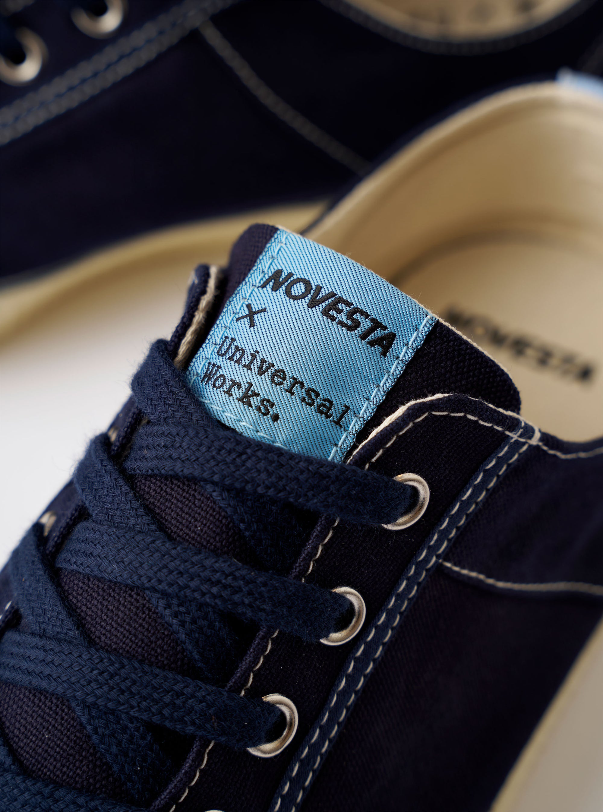 The Novesta for Universal Works Navy/Pale Blue Canvas Star Master is shown from a side profile against a white background. The low-top sneaker features a navy cotton canvas upper with contrasting pale blue stitching and silver-tone metal eyelets. It has a chunky, textured ecru rubber sole and matching rubber toe cap. A dual-branded woven tag is visible on the tongue, and