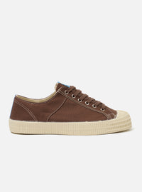 The Novesta for Universal Works Brown/Pale Blue Canvas Star Master is shown from a side profile as a flat lay against a white background. The low-top sneaker features a brown cotton canvas upper with contrast stitching and metallic eyelets for the laces. A signature chunky, vulcanized ecru rubber sole and toe cap are prominent, showing a distinct tire-like texture on the sides. A pale blue pull tab is attached to the heel.