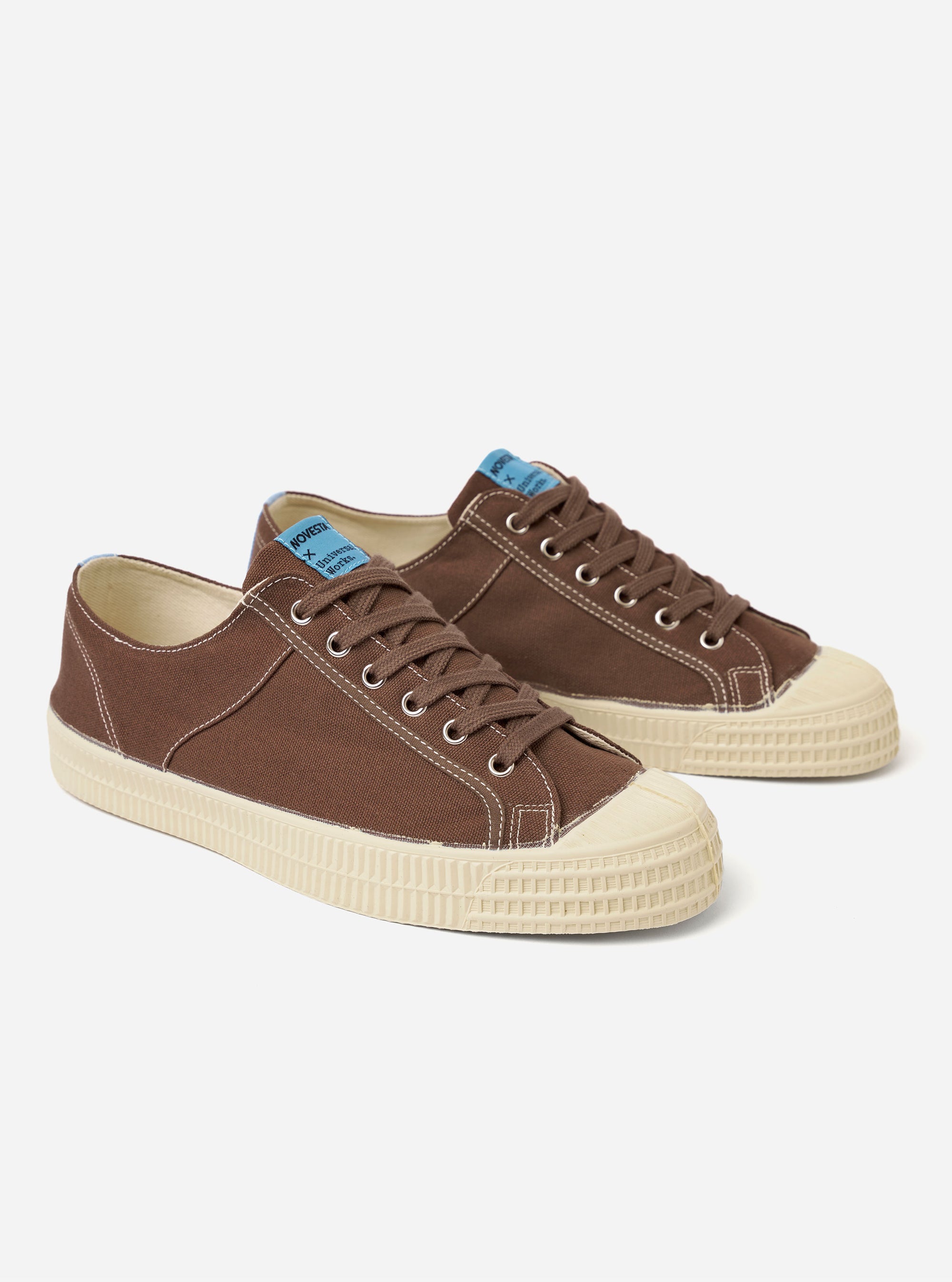The Novesta for Universal Works Brown/Pale Blue Canvas Star Master is shown from a side profile view against a white background. The sneaker features a brown cotton canvas upper with contrast stitching, metallic eyelets, and a signature chunky ecru vulcanized rubber sole. A pale blue pull tab is prominent on the heel, and a dual-branded woven tag is visible on the tongue.