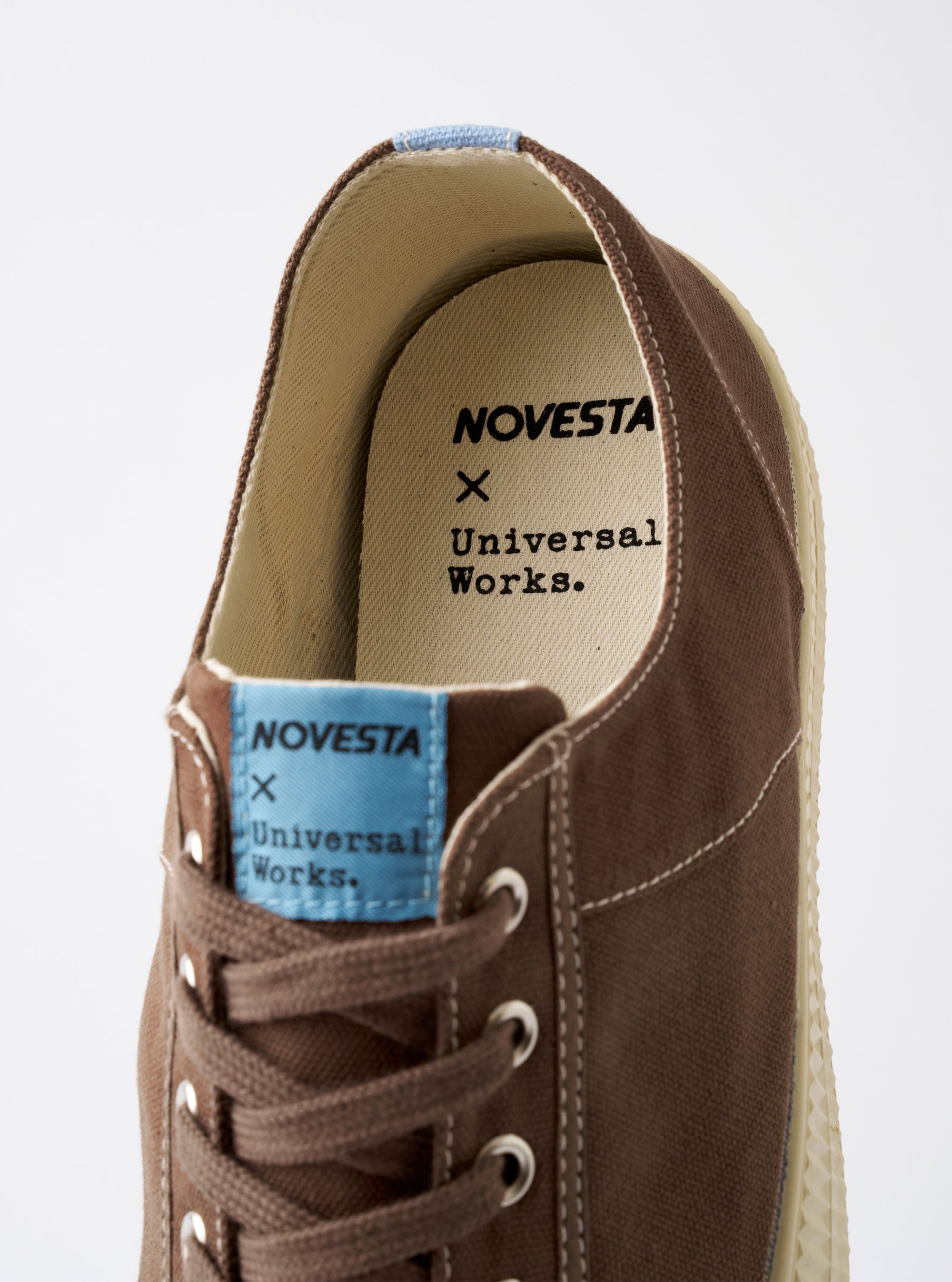 The Novesta for Universal Works Brown/Pale Blue Canvas Star Master shoe is shown from a side profile against a plain white background. The sneaker features a brown cotton canvas upper with contrast stitching, silver metallic eyelets, and tonal brown laces. It has a chunky ecru-colored vulcanized rubber sole with a matching rubber toe cap and a signature pale blue pull tab at the heel.