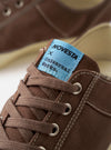 The Novesta for Universal Works Brown/Pale Blue Canvas Star Master sneaker is shown from a side profile against a white background. The shoe features a dark brown cotton canvas upper with contrast stitching, silver metallic eyelets, and a chunky ecru-colored vulcanized rubber sole with a textured toe cap. A distinctive pale blue pull tab is visible on the heel, and a dual-branded woven tag is stitched onto the tongue.