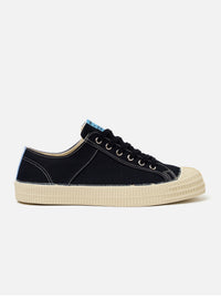 The Novesta for Universal Works Black/Pale Blue Canvas Star Master is shown from a side profile as a flat lay against a white background. The low-top sneaker features a black cotton canvas upper with contrast white stitching and metallic eyelets. A signature chunky ecru vulcanized rubber sole and toe cap are prominent, with a pale blue flash detail on the heel.