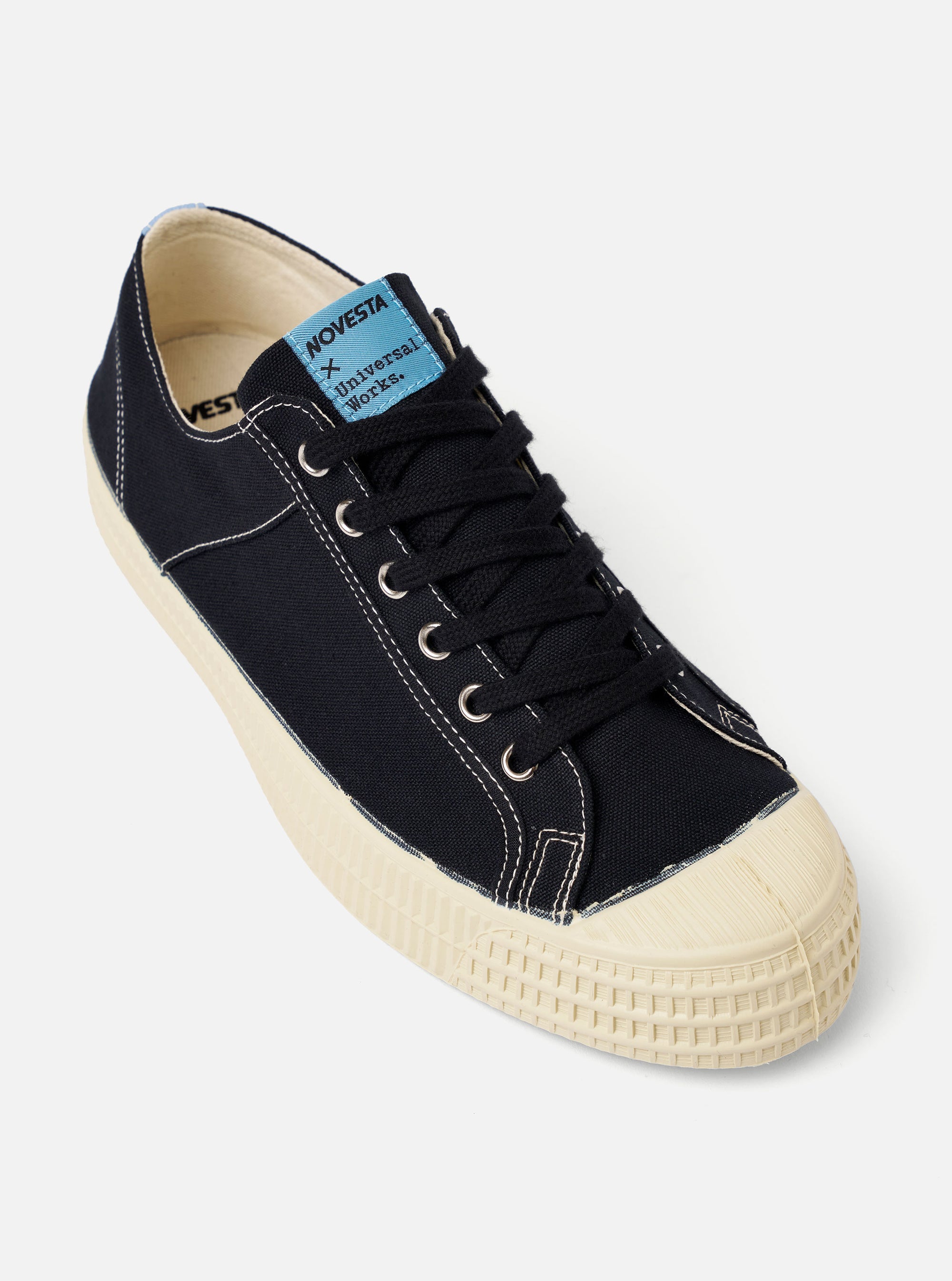 The Novesta for Universal Works Black/Pale Blue Canvas Star Master is shown from a side profile view against a white background. The sneaker features a black cotton canvas upper with contrast stitching, silver metallic eyelets, and black laces. A prominent pale blue pull tab is located at the heel, and a dual-branded woven tag is visible on the tongue. The shoe is built on a chunky, textured ecru vulcanized rubber sole with a matching rubber shell toe.