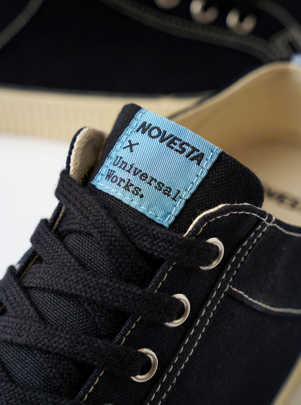 The Novesta for Universal Works Black/Pale Blue Canvas Star Master is shown from a high-angle, top-down perspective against a white background. The low-top sneaker features a black cotton canvas upper with contrast white stitching, metallic eyelets, and black laces. A prominent feature is the light blue heel tab and the dual-branded woven logo on the tongue. The chunky, vulcanized rubber sole is ecru with a textured toe cap.