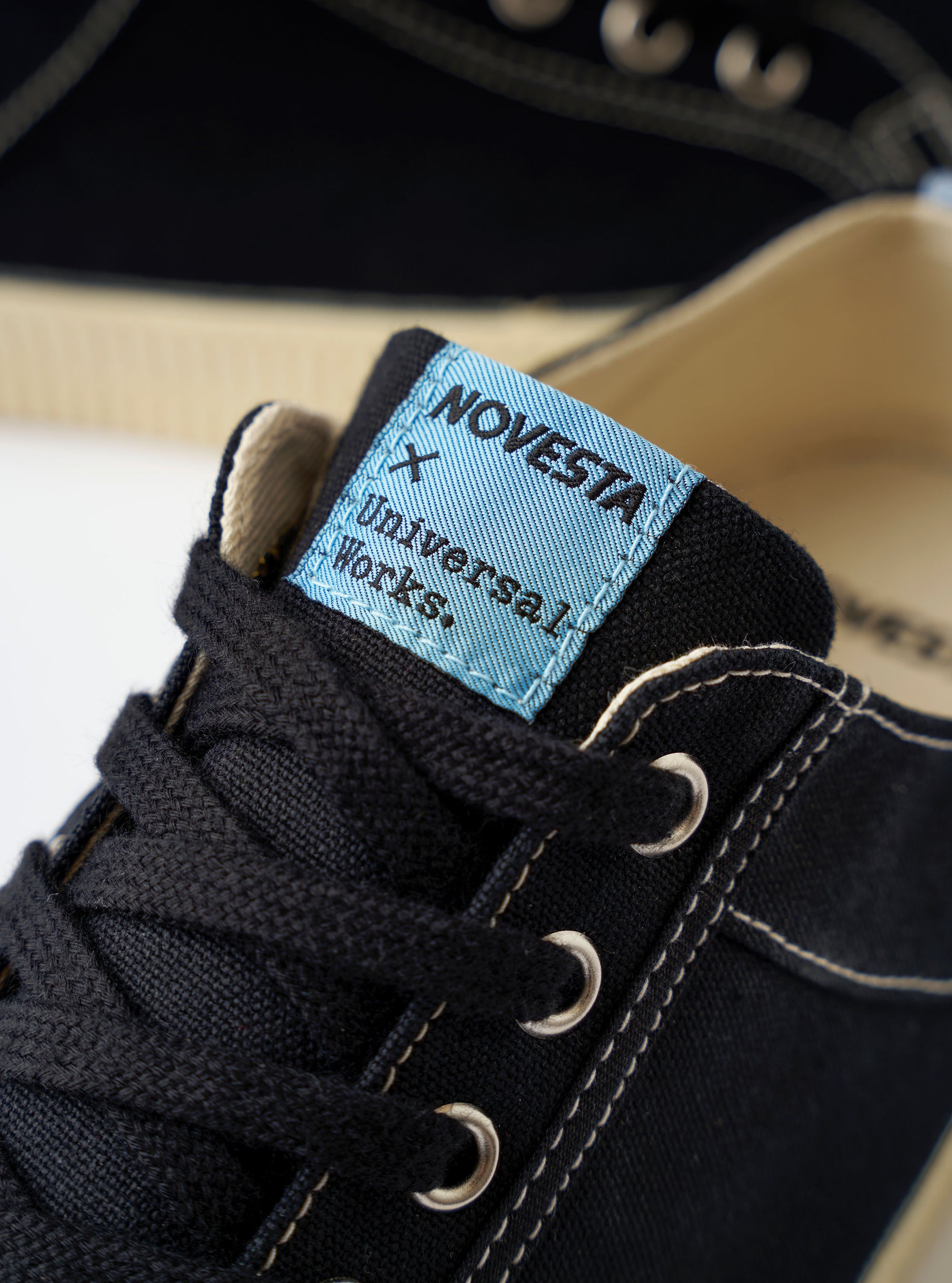 The Novesta for Universal Works Black/Pale Blue Canvas Star Master is shown from a high-angle, top-down perspective against a white background. The low-top sneaker features a black cotton canvas upper with contrast white stitching, metallic eyelets, and black laces. A prominent feature is the light blue heel tab and the dual-branded woven logo on the tongue. The chunky, vulcanized rubber sole is ecru with a textured toe cap.