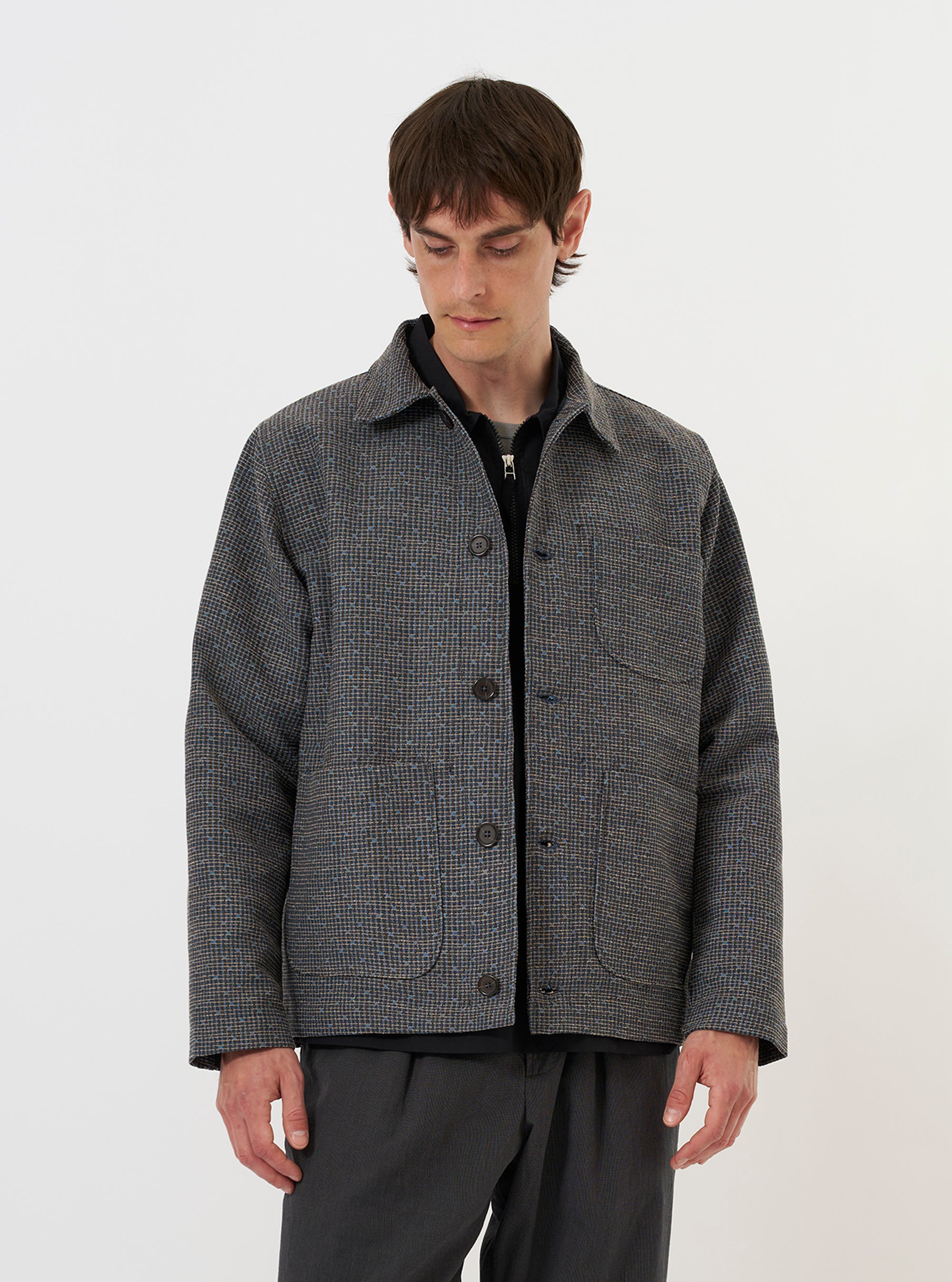 A model is wearing the Grey/Blue Jacquard Weave Field Jacket against a plain white background. The jacket features a complex textured pattern in shades of grey and blue, a classic pointed collar, a button-down front, and several large utility patch pockets. The model is shown from the waist up, highlighting the garment's relaxed fit and structured design. No mannequin is present in the image.