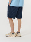 A model is wearing the Navy Chester Stripe Pleated Track Short against a plain white background. The navy blue shorts feature a thin white vertical stripe pattern and a pleated front. The image captures the model from the waist down, highlighting the relaxed fit and the side profile of the garment. The model is standing and is also wearing dark-colored shoes and white socks.