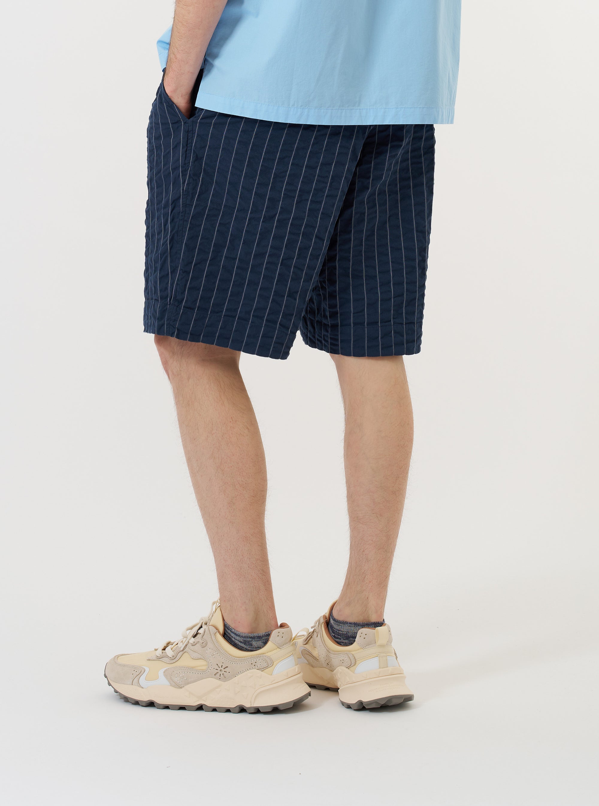 A model is wearing the Navy Chester Stripe Pleated Track Short against a plain white background. The navy blue shorts feature a thin white vertical stripe pattern and a pleated front. The image captures the model from the waist down, highlighting the relaxed fit and the side profile of the garment. The model is standing and is also wearing dark-colored shoes and white socks.