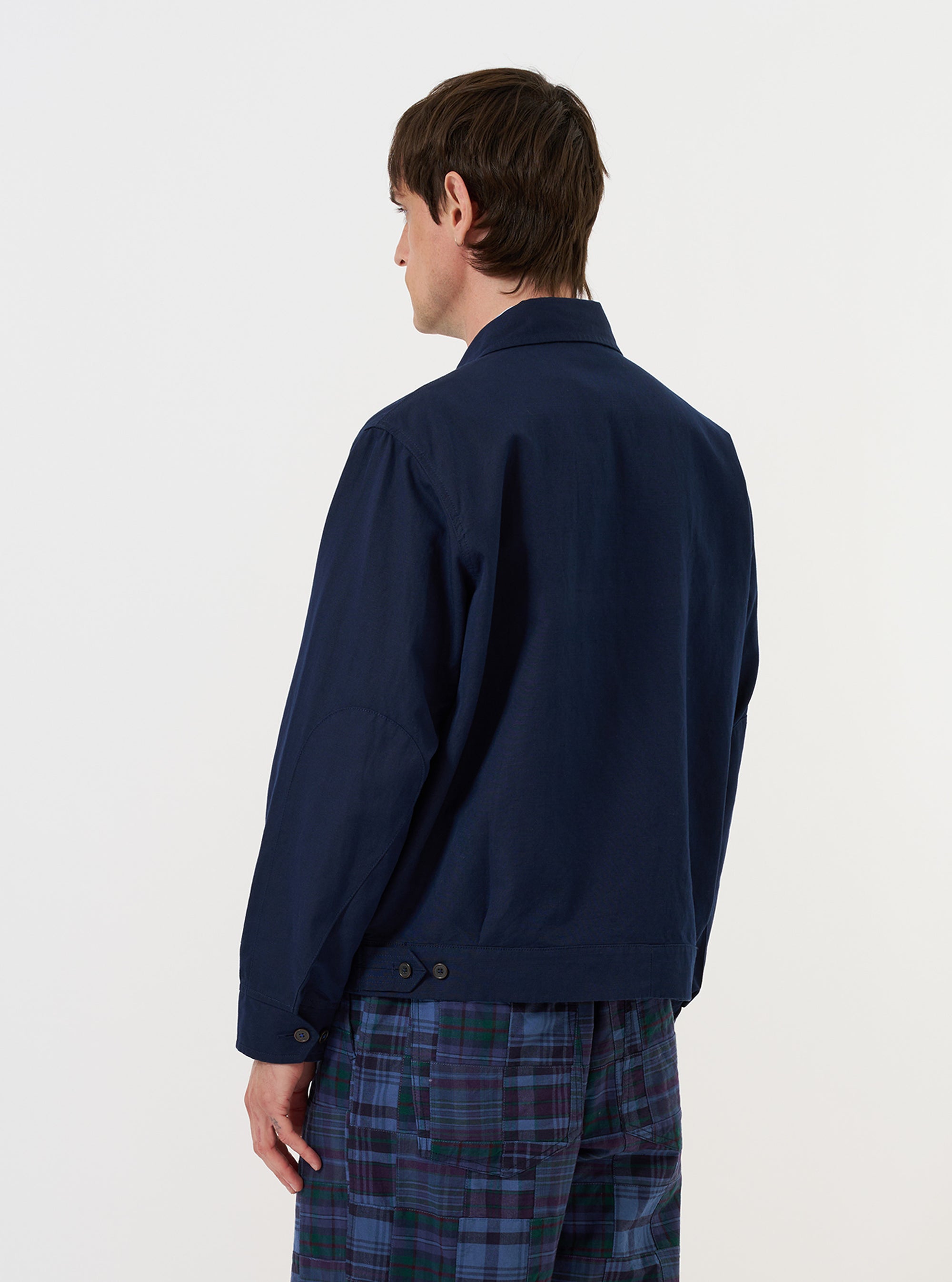 A male model is wearing the Navy Linen Slub Weave E130 Jacket against a plain white background. The model is seen from the side, looking over his shoulder toward the camera, highlighting the jacket's relaxed fit and navy blue textured fabric. The jacket features a classic collar, a silver-toned two-way front zipper, and adjustable buttoned cuffs. He is wearing the jacket over a light-colored top.