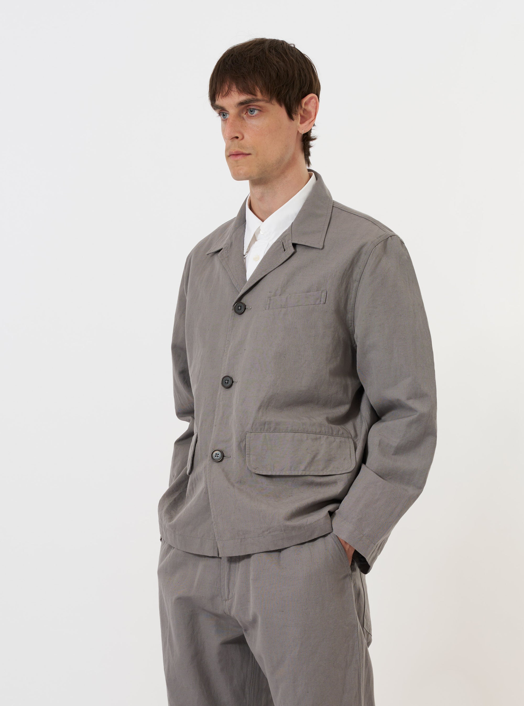 The Mid Grey Linen Slub Weave Franck Jacket is shown laid flat against a solid white background. This grey jacket features a full button front, a classic pointed collar, one chest welt pocket, and two lower flap hip pockets. The image highlights the textured, slubbed weave of the linen-cotton blend fabric.