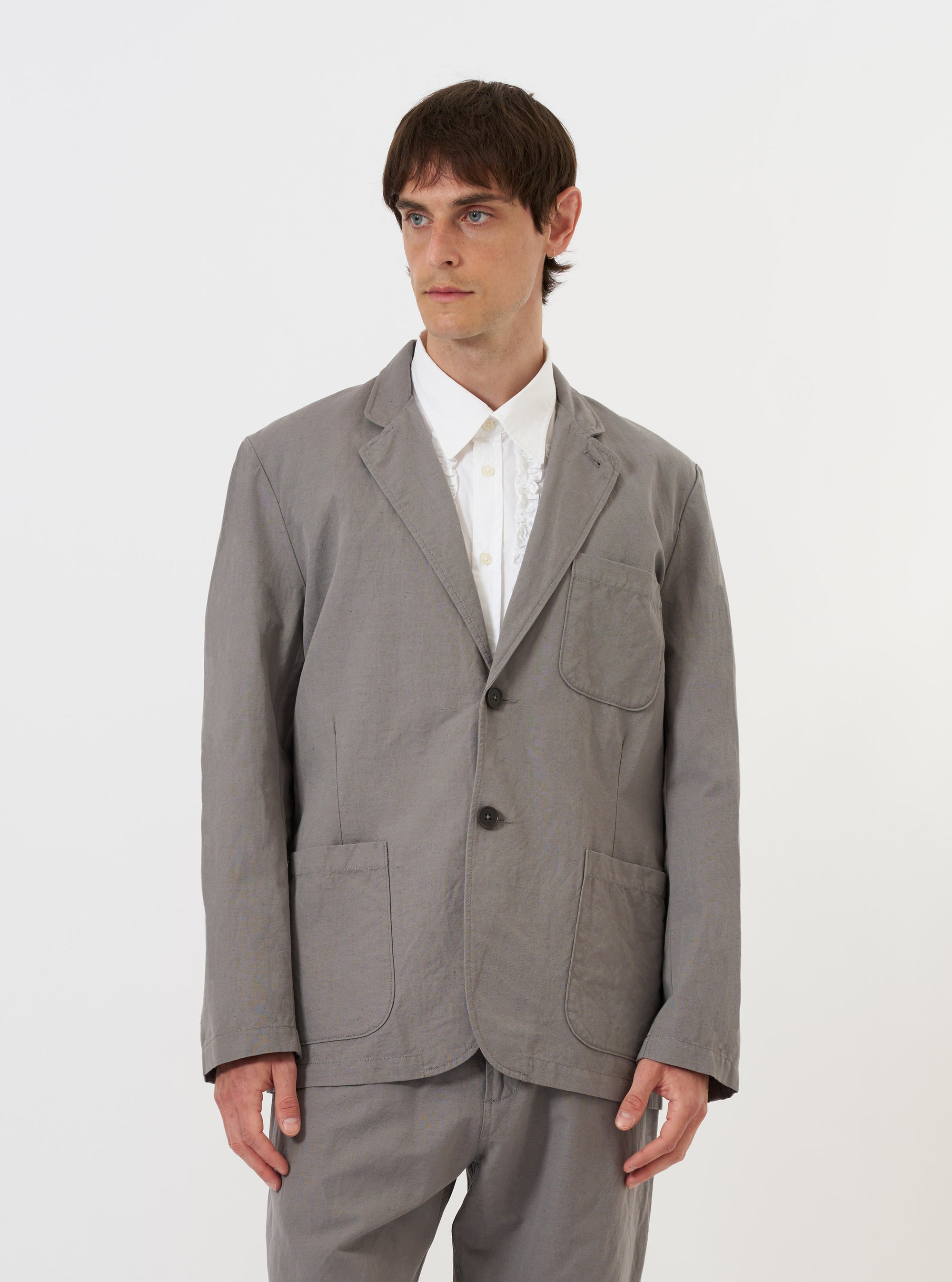 A Mid Grey Linen Slub Weave Two Button Jacket is shown laid flat against a solid white background. The jacket features a relaxed fit with a two-button front, a long and wide notch lapel, two lower patch pockets, and a single chest patch pocket. The fabric has a visible, textured slub weave.