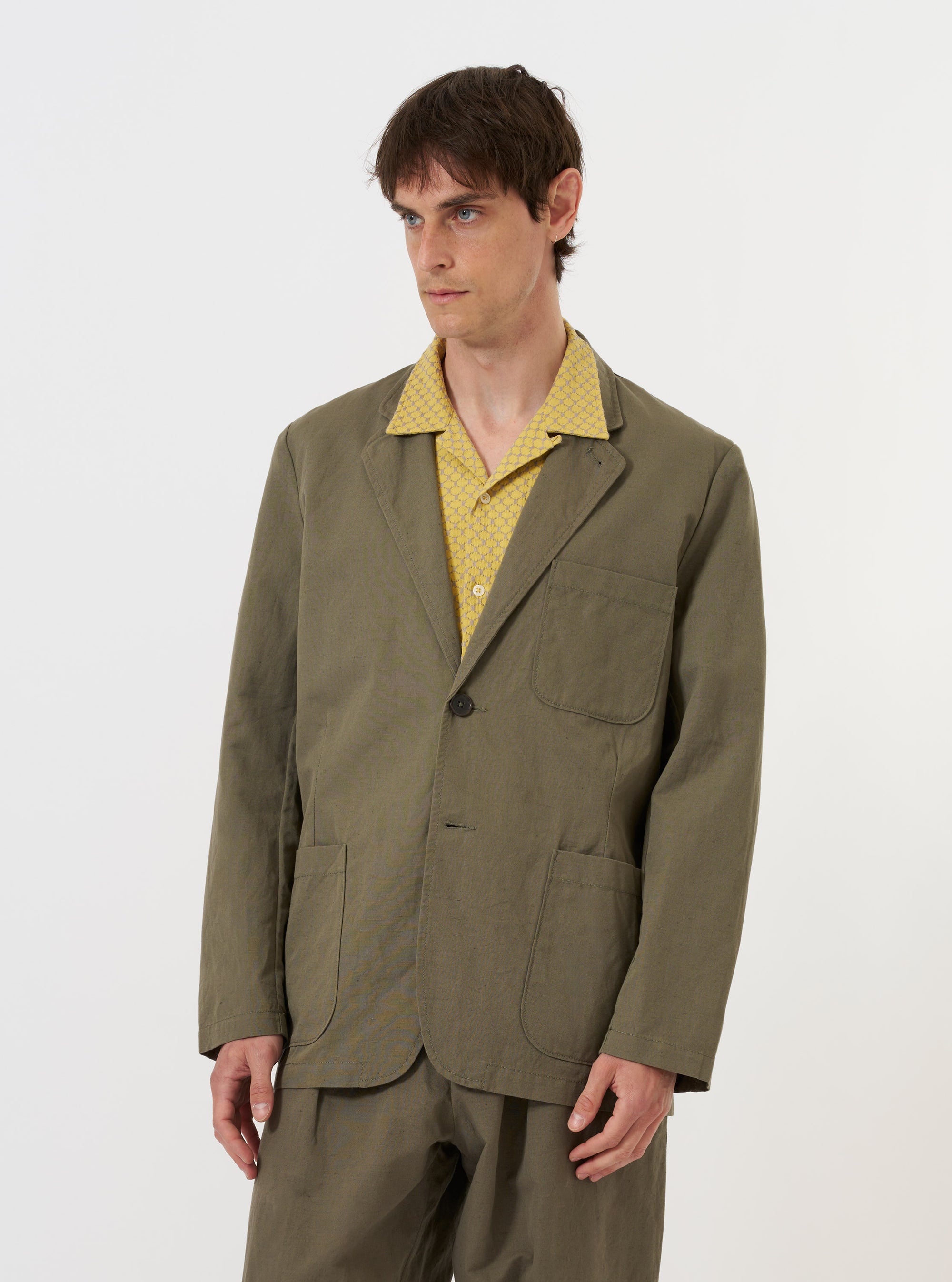 An Olive Linen Slub Weave Two Button Jacket is displayed flat against a plain white background. The garment features a long, wide notch lapel, a two-button front closure, two large lower patch pockets, and a single chest patch pocket. The olive green fabric has a visible textured slub weave.