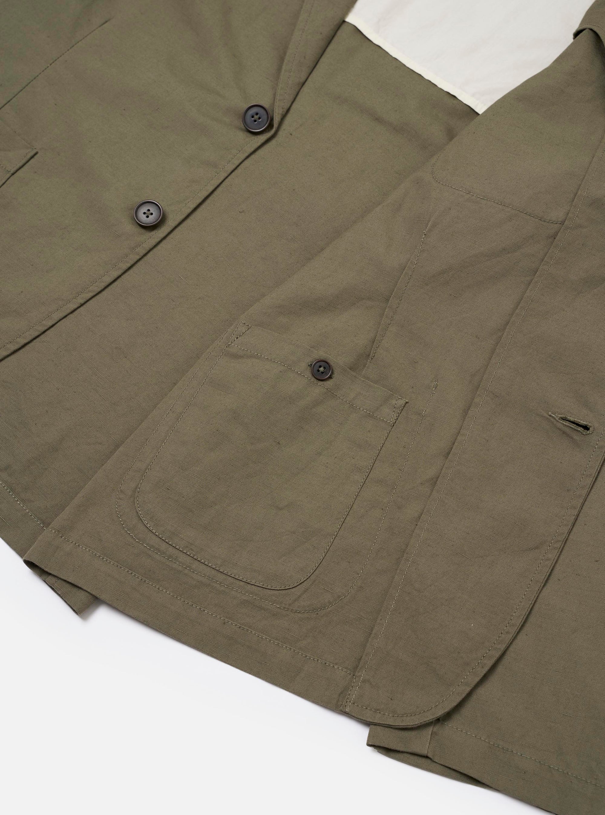 The Olive Linen Slub Weave Two Button Jacket is shown in a close-up detail view, laid flat against a plain white background. The image highlights the textured slub weave of the olive green linen-cotton fabric, focusing on the construction of a straight side flap pocket and the two-button front closure.
