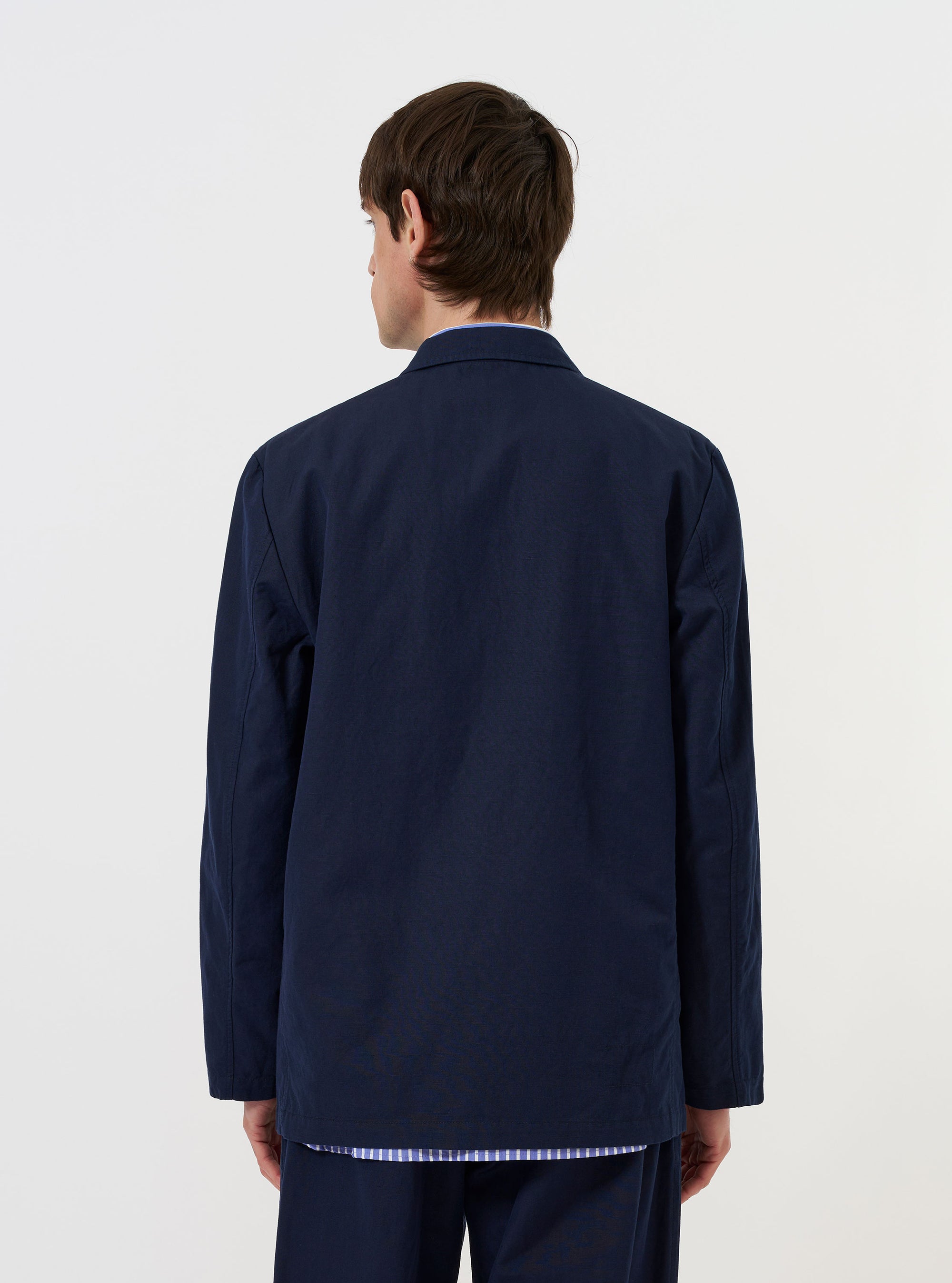 A model is wearing the Navy Linen Slub Weave Two Button Jacket against a plain white background. The image provides a side-rear view of the jacket, highlighting its relaxed fit, unstructured shoulder, and the textured slub weave of the navy linen fabric. The model is standing with their hands by their sides, and no mannequin is present.