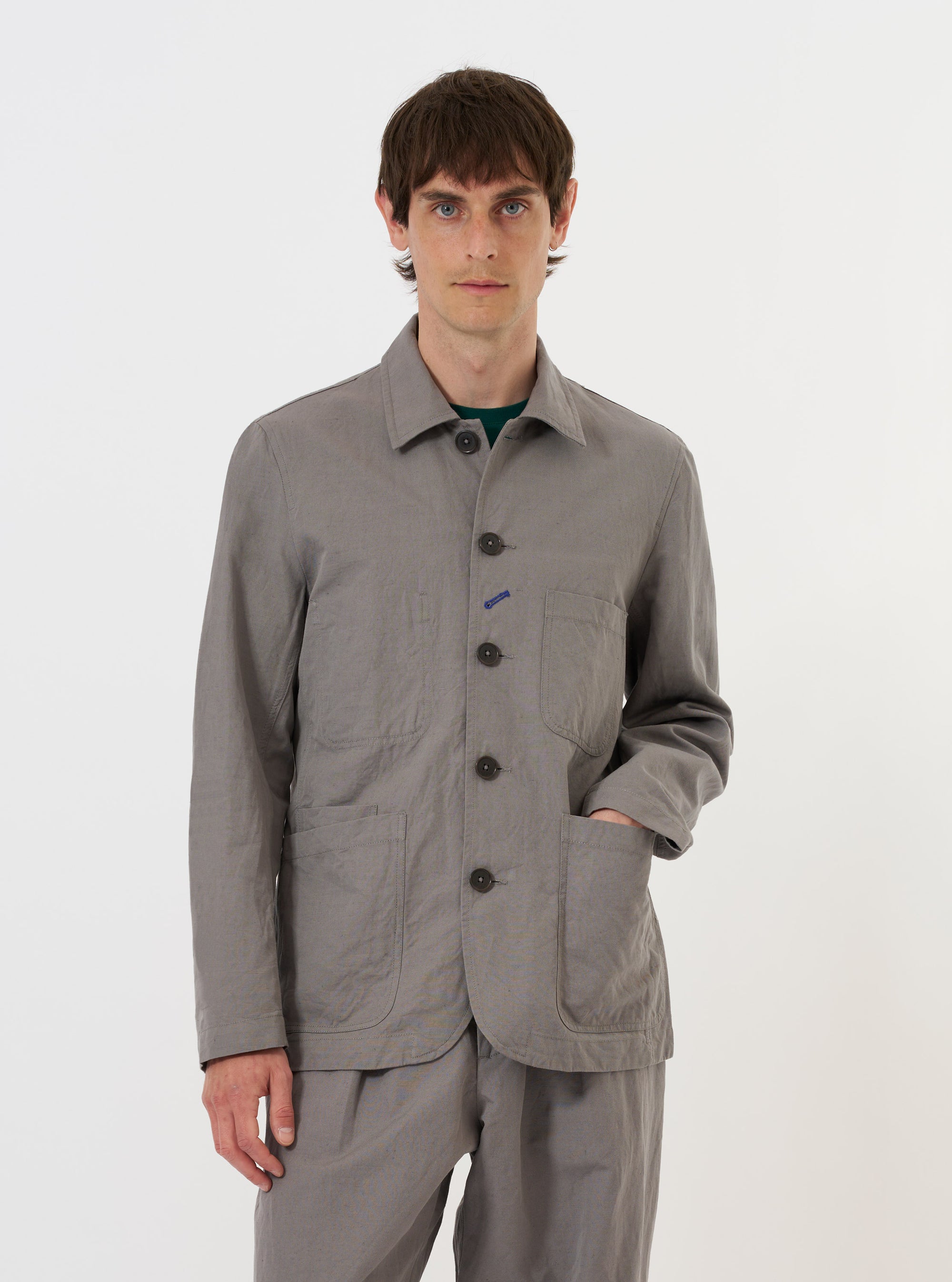 The Mid Grey Linen Slub Weave Bakers Jacket is shown laid flat against a plain white background. This chore-style jacket features a classic pointed collar, a five-button front, and four front patch pockets: one asymmetrical chest pocket and three lower pockets. The mid-grey fabric has a visible slub texture.