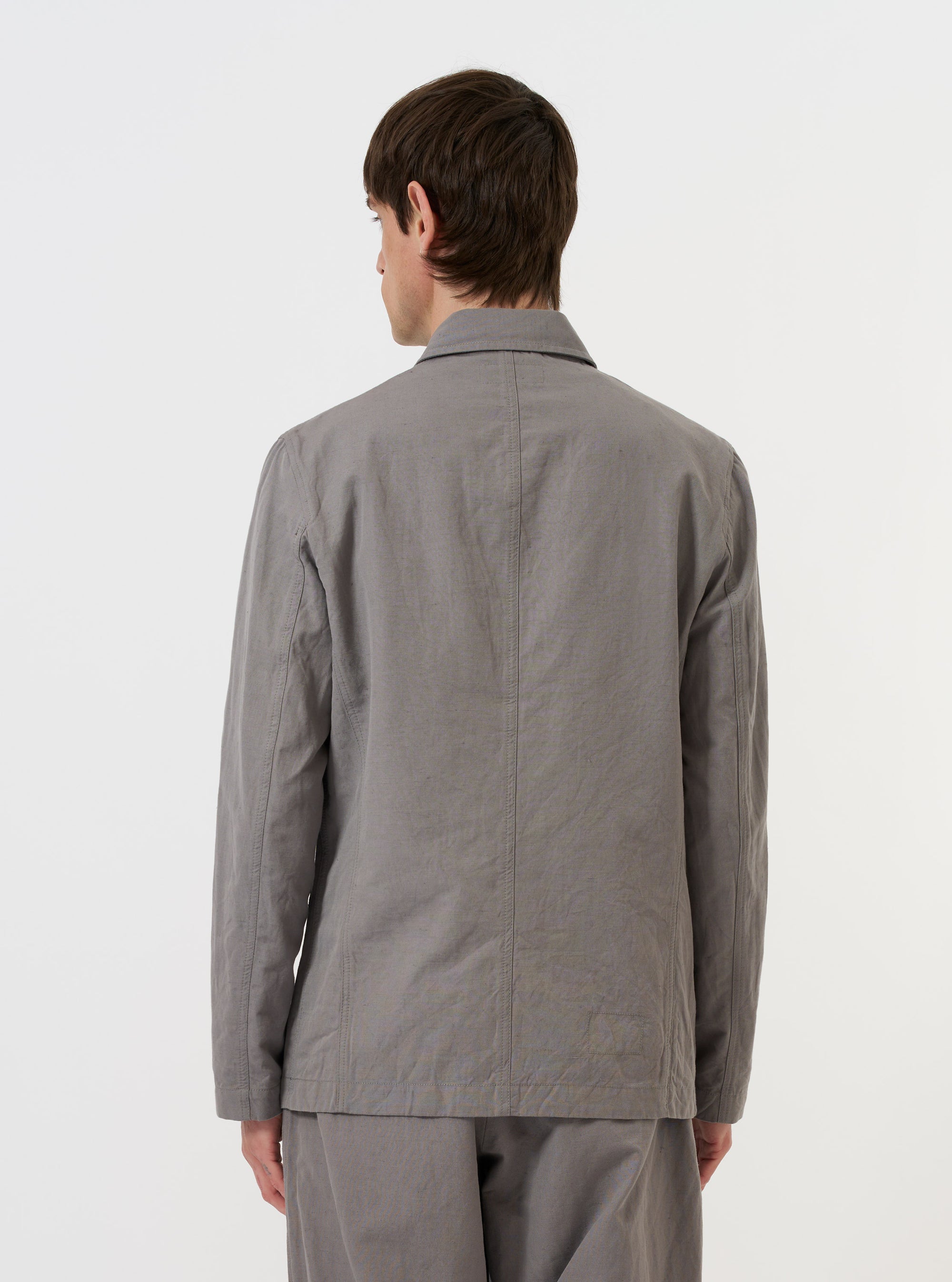 A model is wearing the Mid Grey Linen Slub Weave Bakers Jacket against a plain white background. The image provides a rear view of the jacket, highlighting its straight hem, structured shoulders, and the textured slub weave of the mid-grey fabric. The model is also wearing dark trousers.