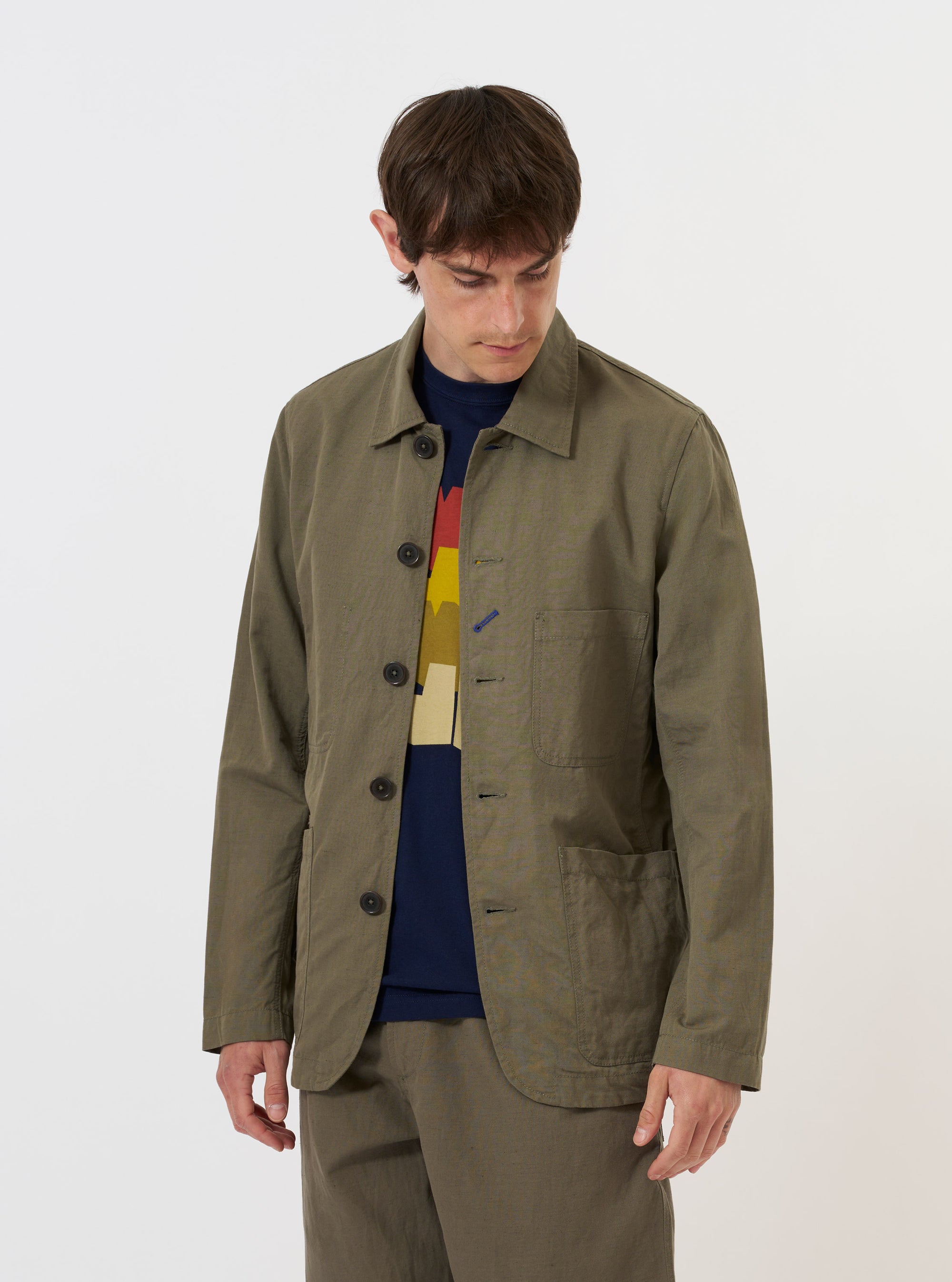 The Olive Linen Slub Weave Bakers Jacket is shown laid flat against a solid white background. The jacket features a classic chore coat silhouette with a five-button front, a pointed collar, and three prominent patch pockets on the front. The olive green fabric has a visible slub texture, and the garment is finished with a straight hem.