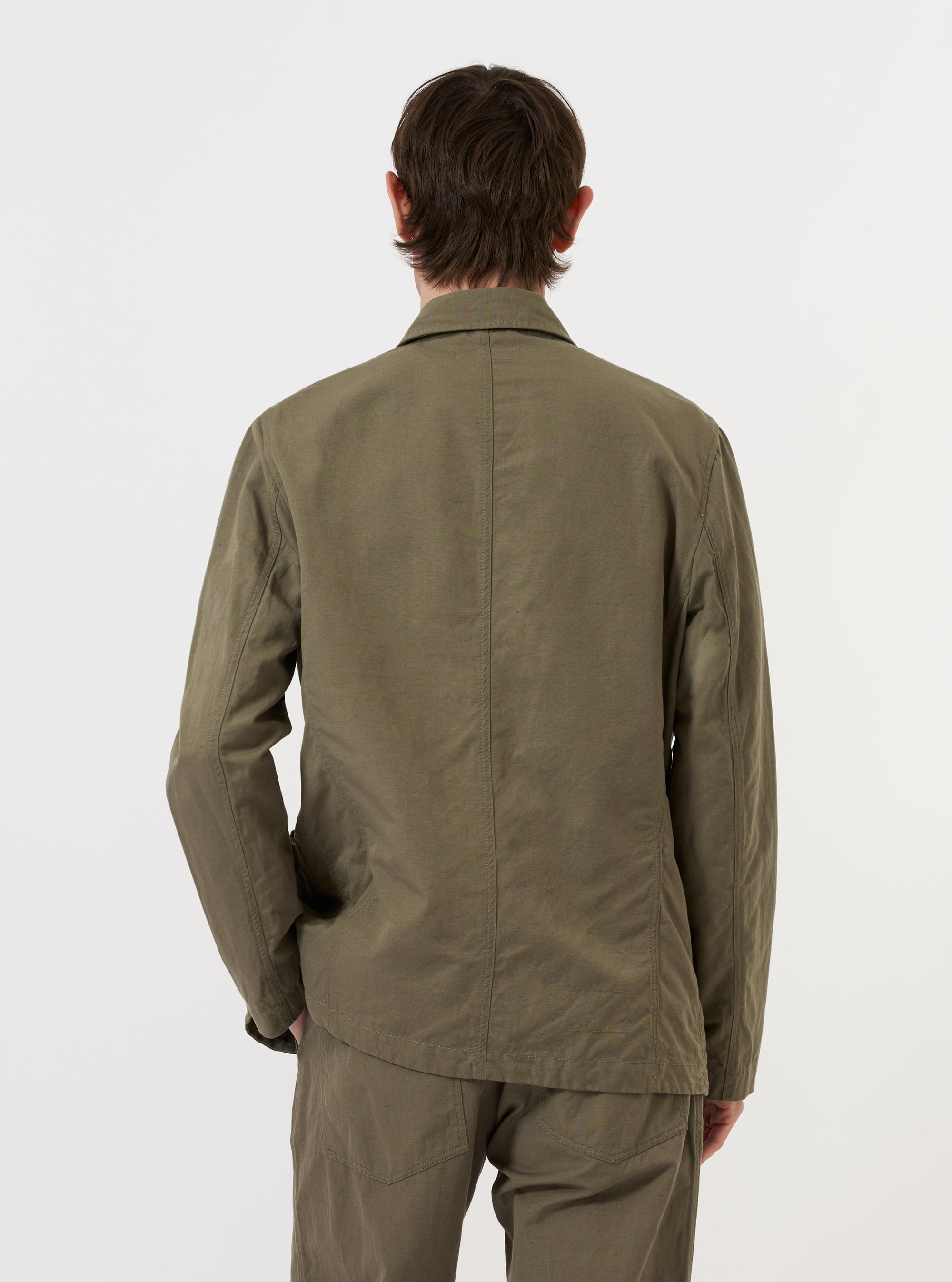 The Olive Linen Slub Weave Bakers Jacket is shown from the back, laid flat against a solid white background. The image highlights the jacket's straight hem, central seam, and the textured slub weave of the olive green linen-cotton fabric.